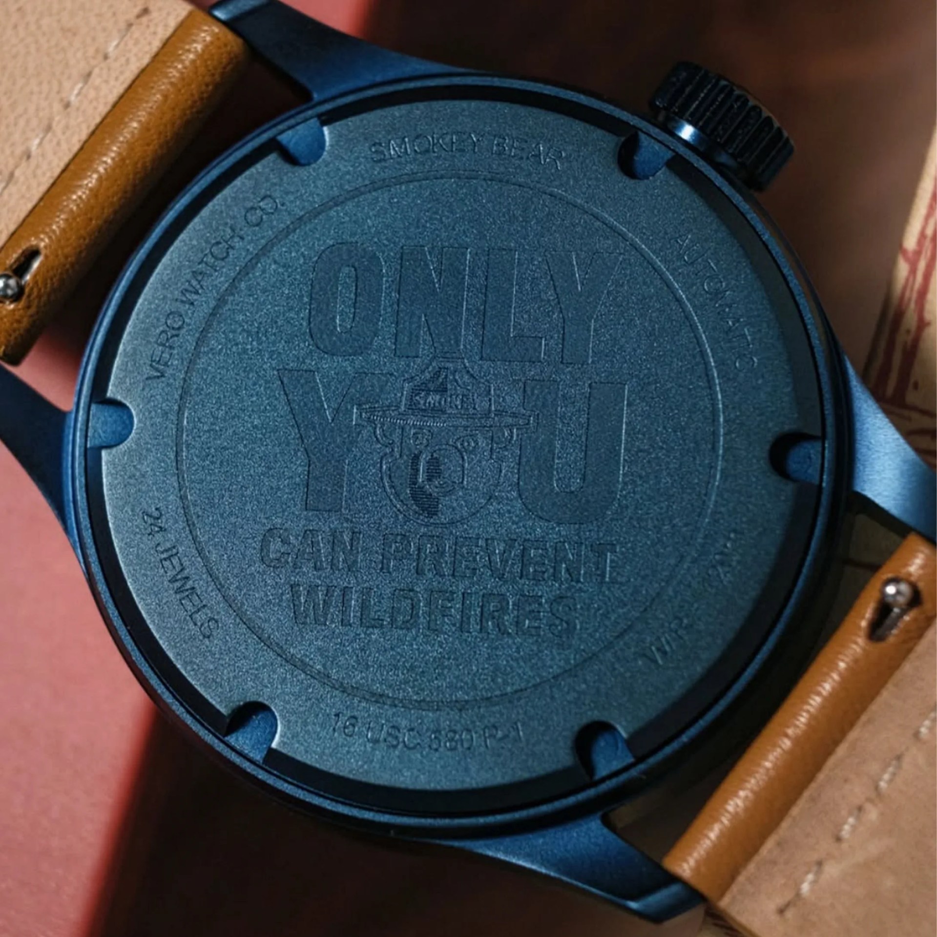 Close-up of a black watch back with engraved text "ONLY YOU CAN PREVENT WILDFIRES" and a brown leather strap.