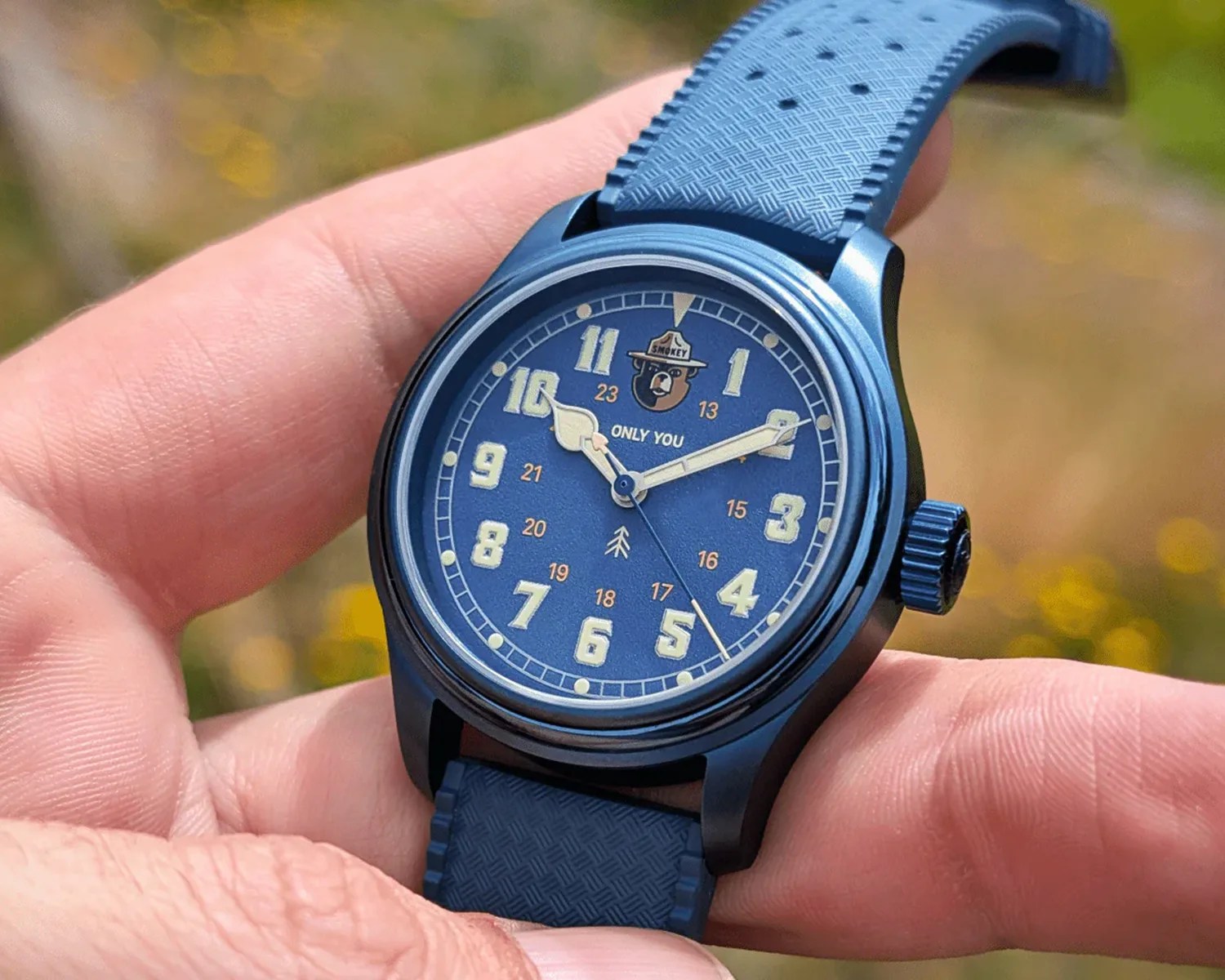 Blue wristwatch with textured strap, large luminous numbers, and a Smokey Bear logo on the dial held in a hand.