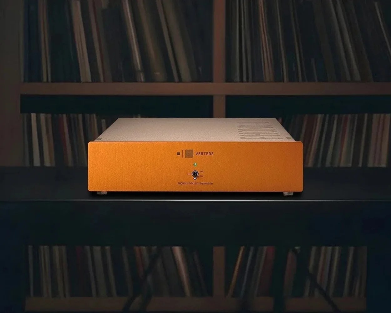 Orange and silver Vertere PHONO-1 MM/MC preamplifier on a black reflective surface with a bookshelf in the background.