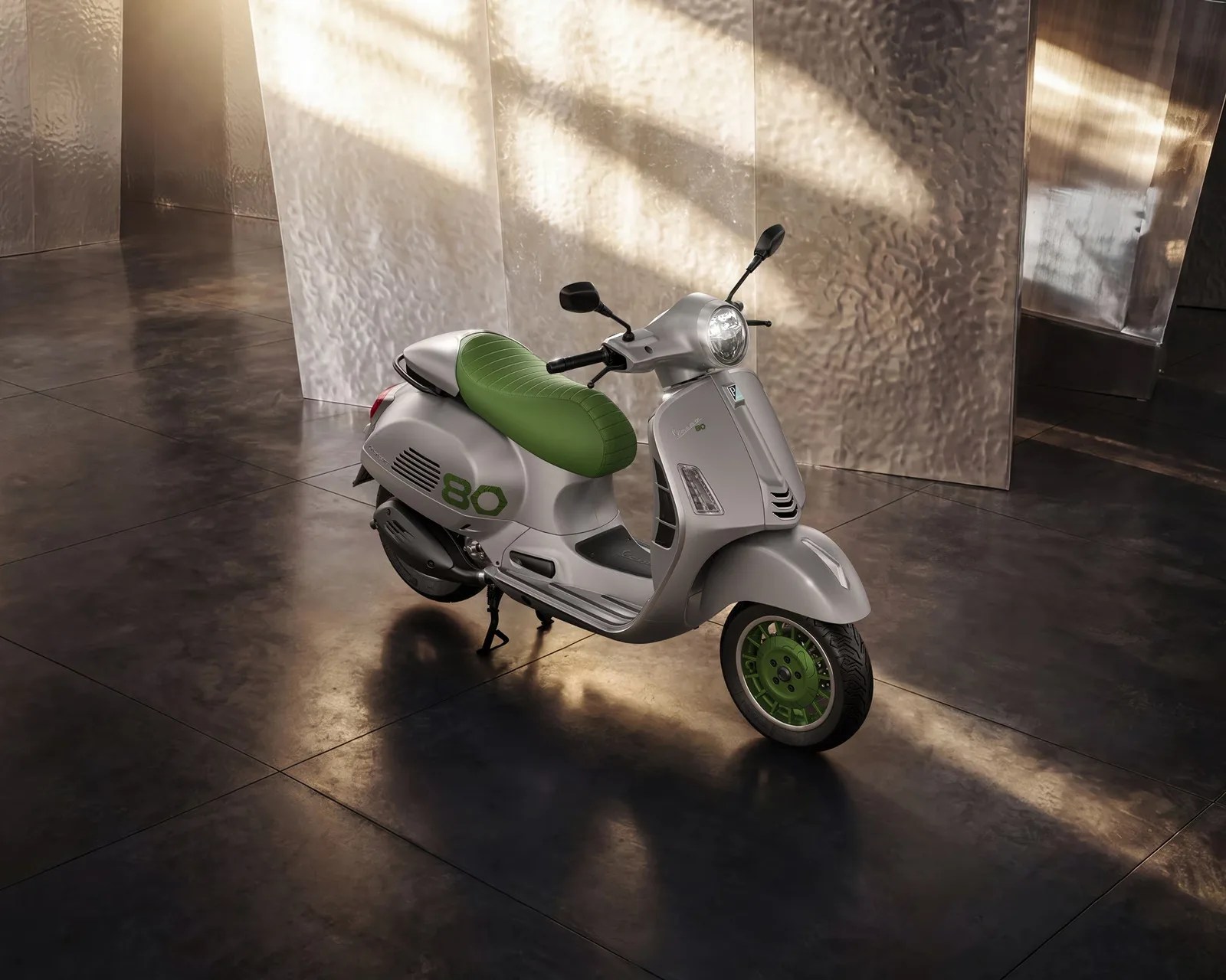 Silver Vespa scooter with green seat and green wheel rims parked indoors on a dark tiled floor.
