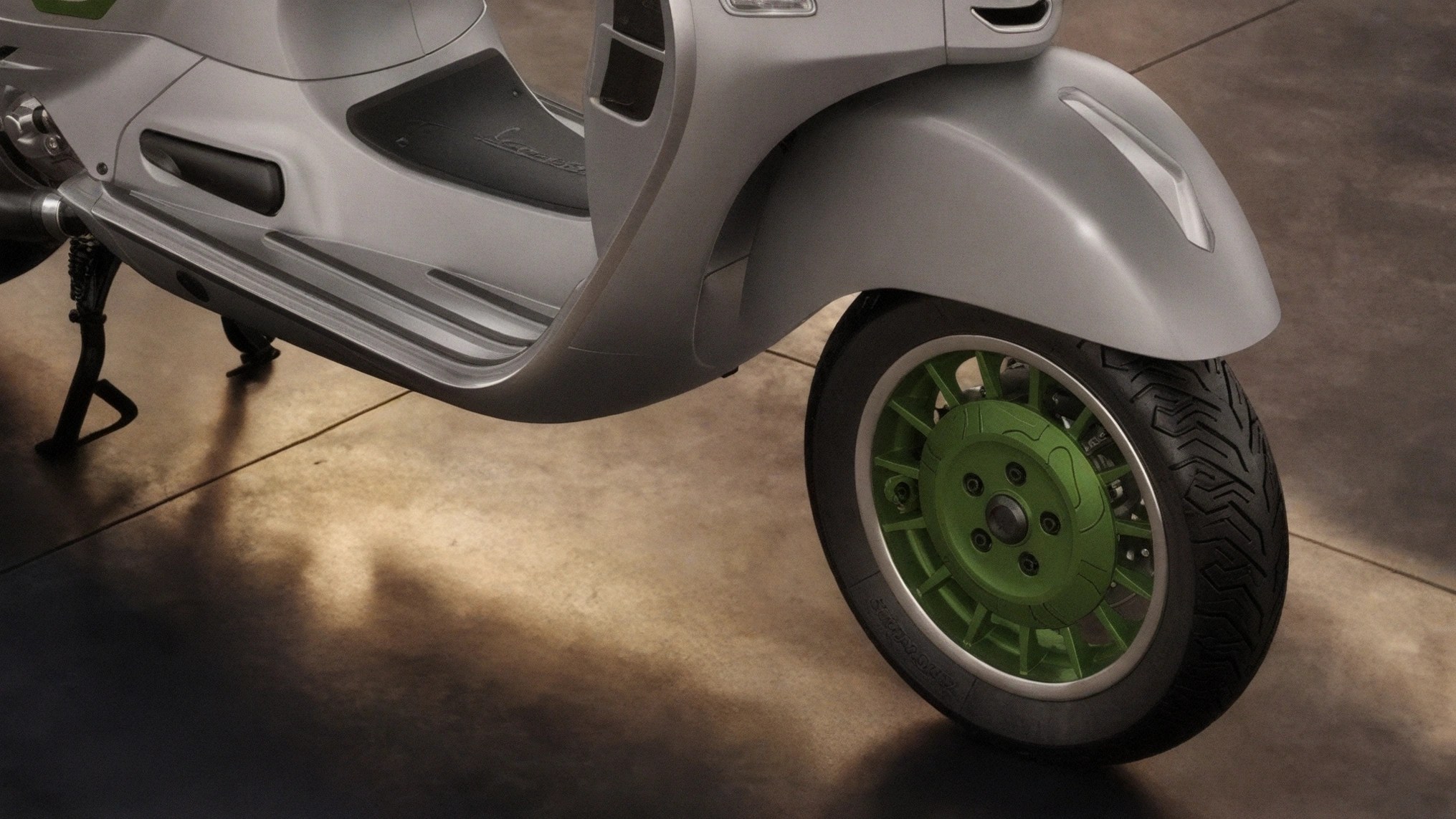 Close-up of a silver scooter's front wheel with a green hub and black tire on a concrete floor.