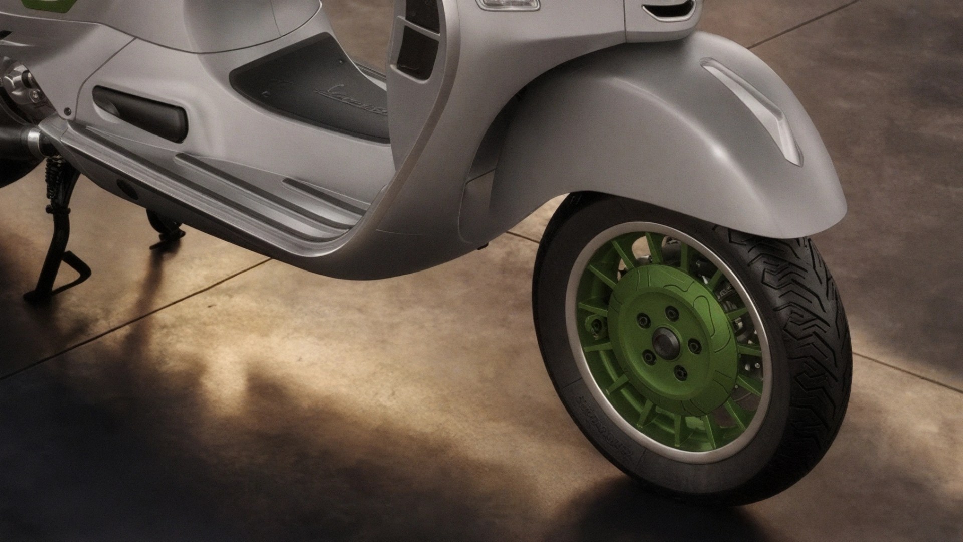 Close-up of a silver scooter's front wheel with a green hub and black tire on a concrete floor.