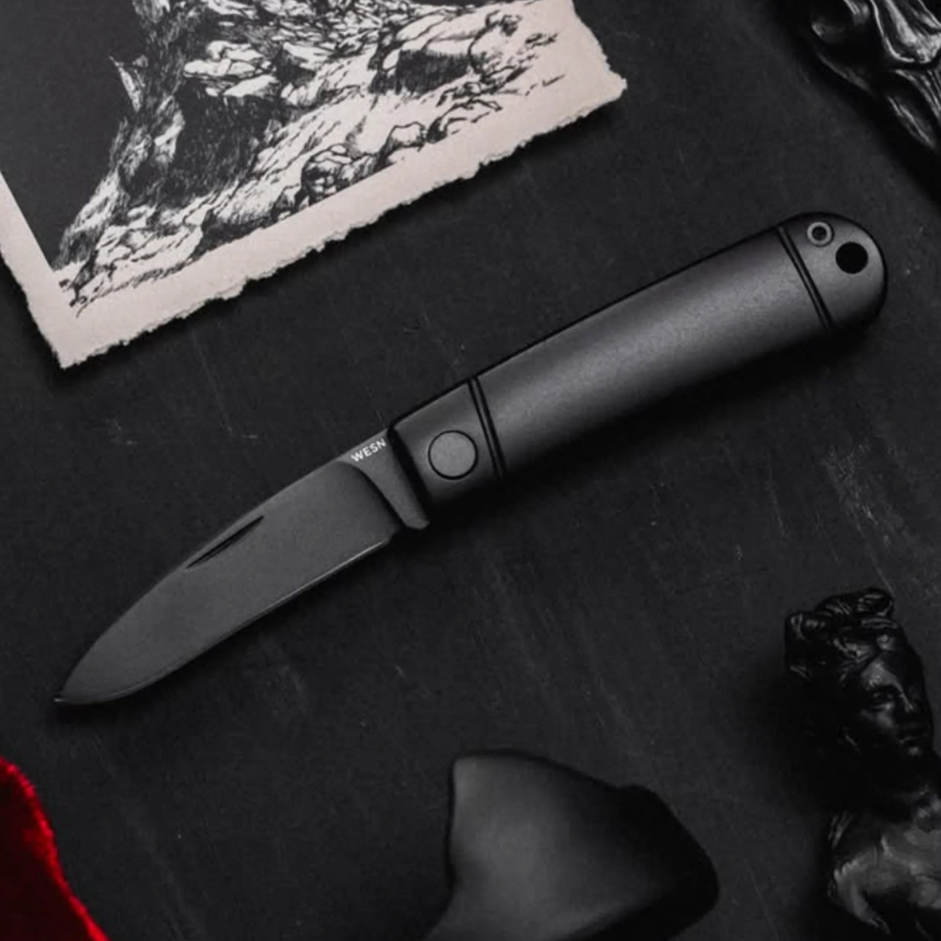 Black folding knife with a matte finish handle on a dark surface next to a torn paper with a sketch.