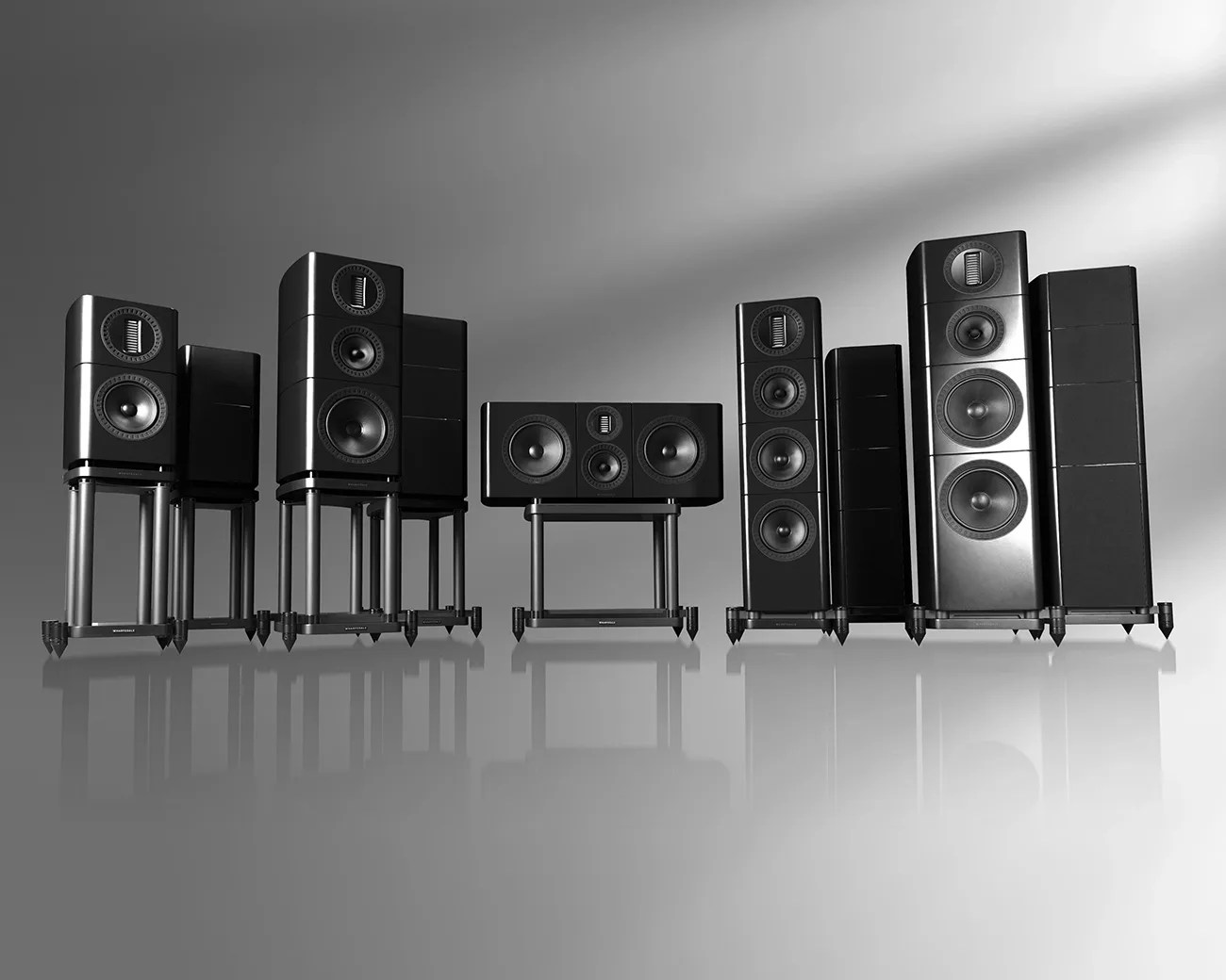 Set of six black high-end speakers of varying sizes on metal stands, arranged in a row on a reflective surface.
