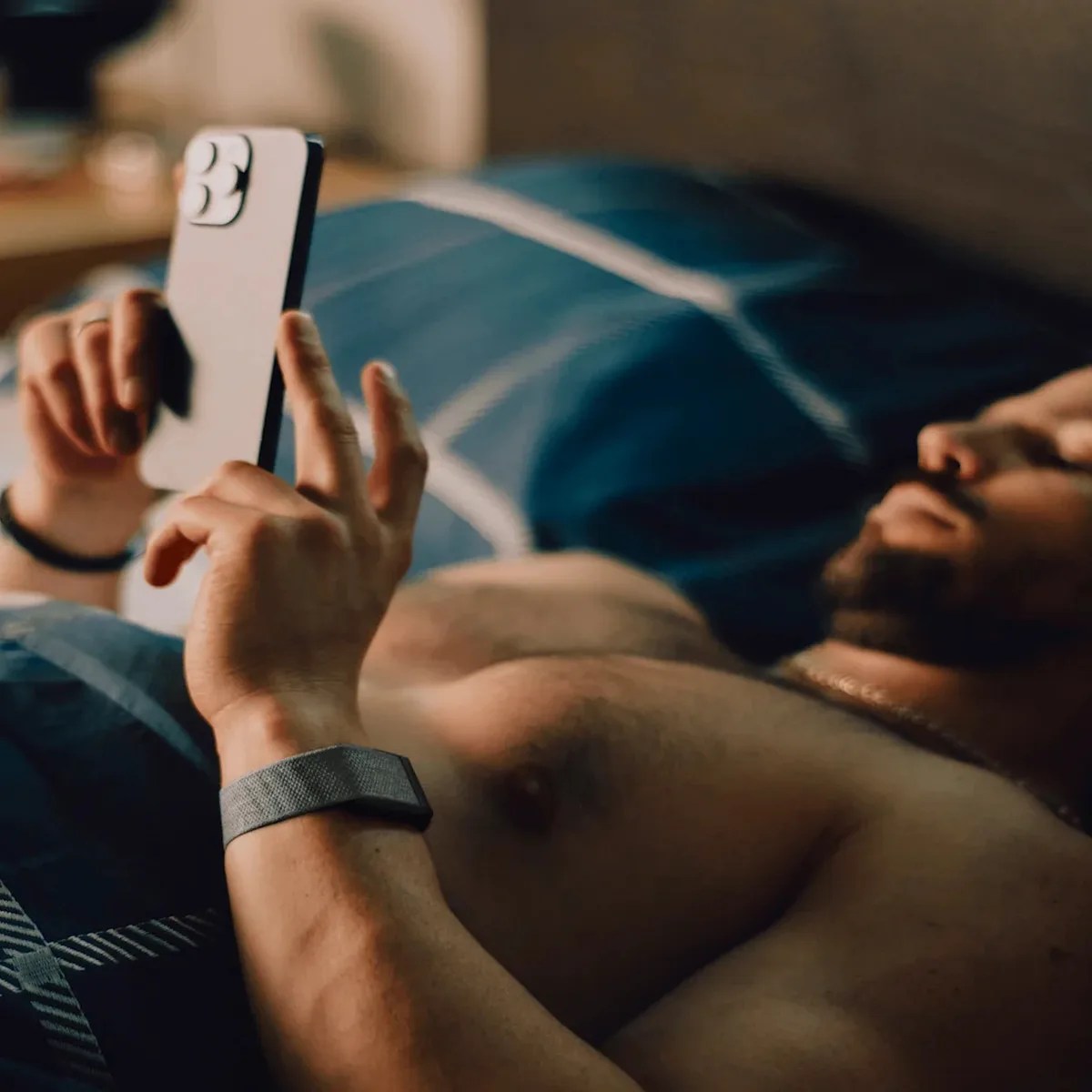 Person lying shirtless on a bed with blue and white bedding, holding a white smartphone above them.