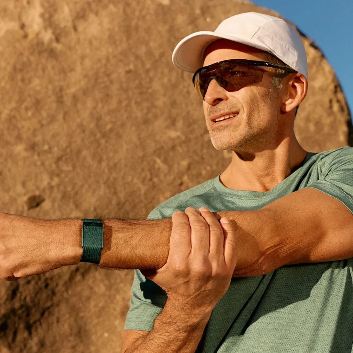 Man in a white cap and sunglasses stretching his arm outdoors wearing a green fitness tracker on his wrist.