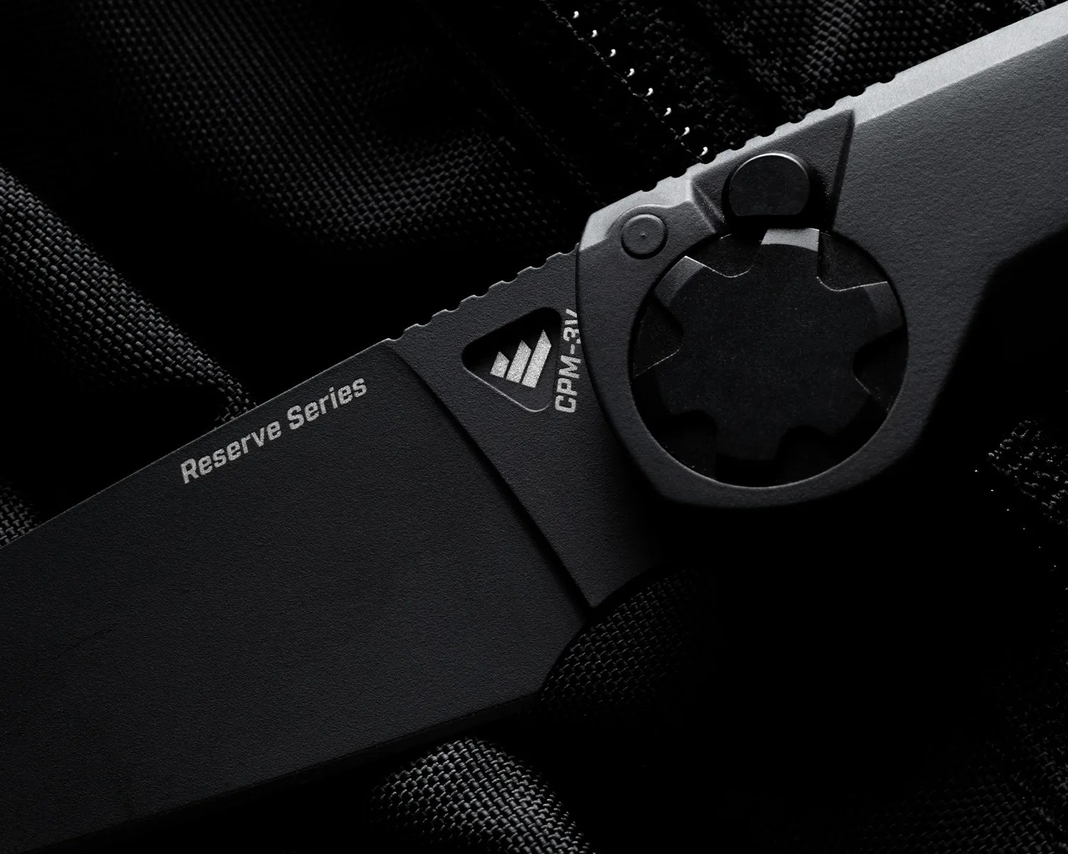 Close-up of a black folding knife blade with "Reserve Series" and "CPM-3V" markings on a textured black fabric background.