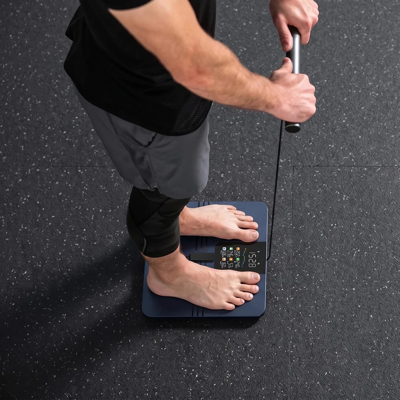 Person standing barefoot on a digital body composition scale holding a connected handle with a cable.