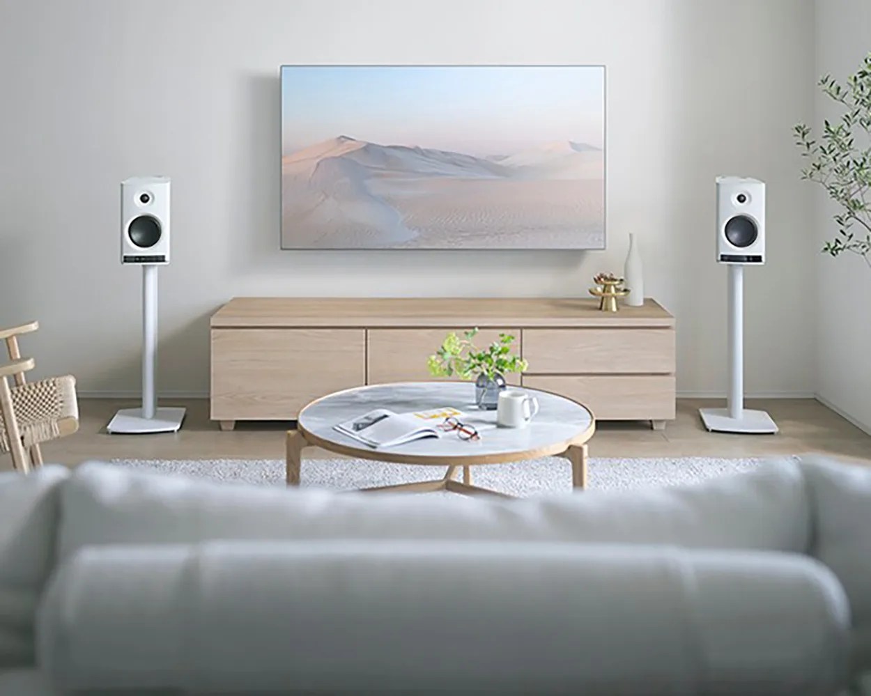 Living room with light wood media console, wall-mounted TV showing desert scene, white floor-standing speakers, round marble-top coffee table, and gray sofa in foreground.