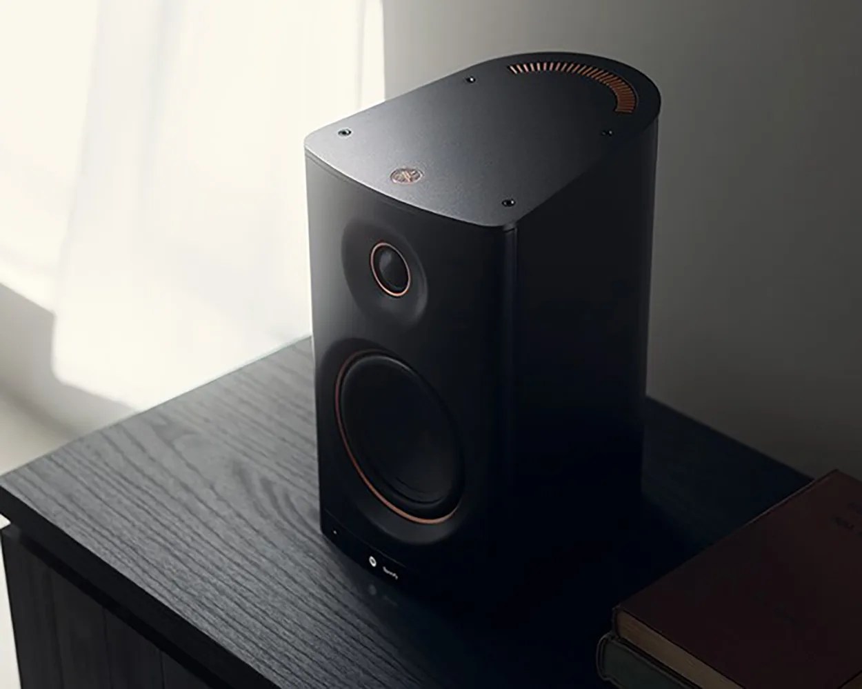 Black rectangular speaker with copper accents on a dark wood surface next to stacked books.