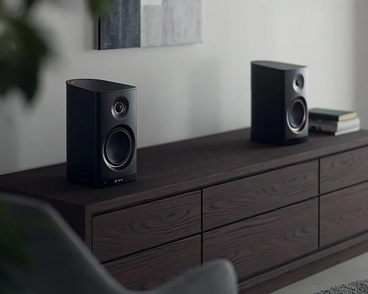 Pair of black bookshelf speakers on a dark wooden cabinet with stacked books on one side.