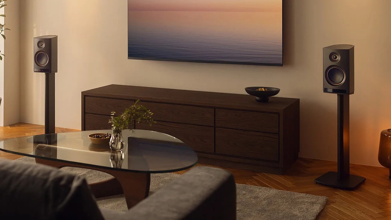 Pair of black bookshelf speakers on stands flanking a dark wood media console with a glass coffee table in front.