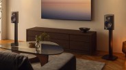 Pair of black bookshelf speakers on stands flanking a dark wood media console with a glass coffee table in front.