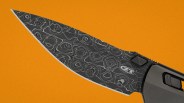 Close-up of a black Damascus steel knife blade with a patterned texture and a partially visible gray handle on an orange background.