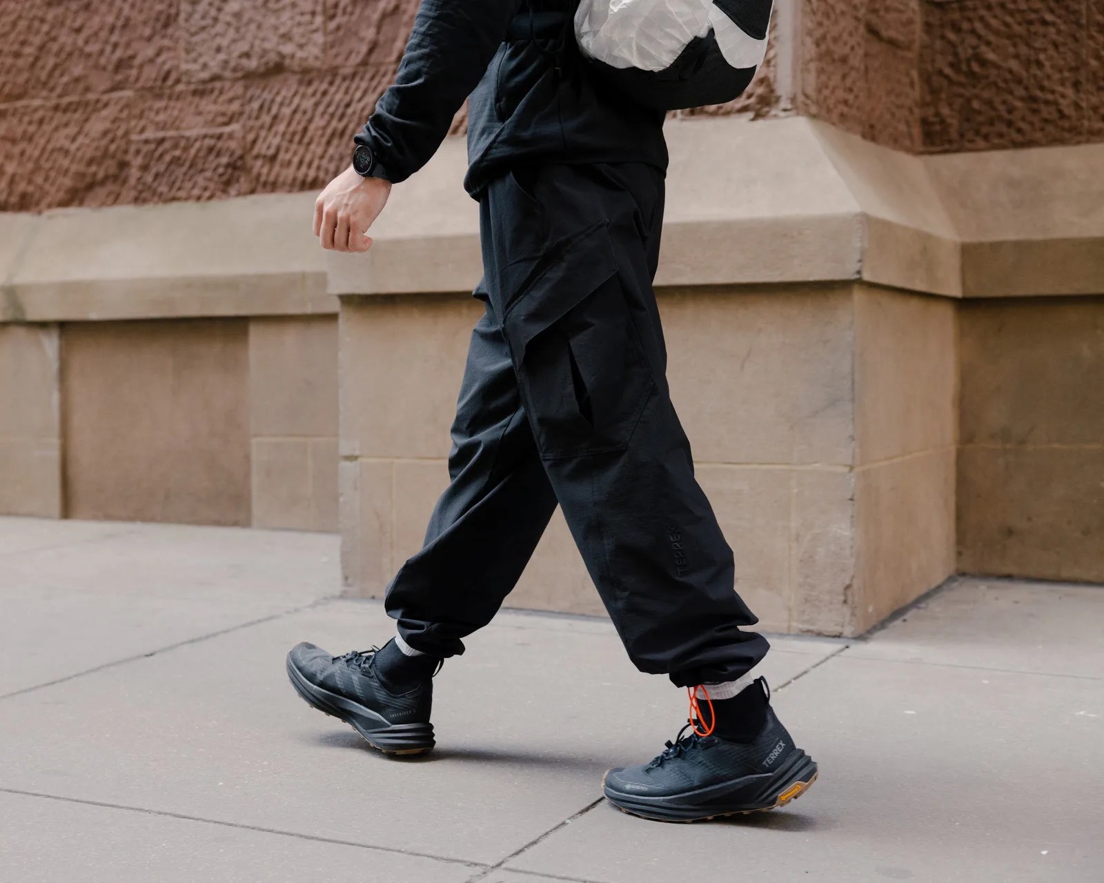 Person wearing black cargo pants and black Terrex sneakers walking on a city sidewalk.