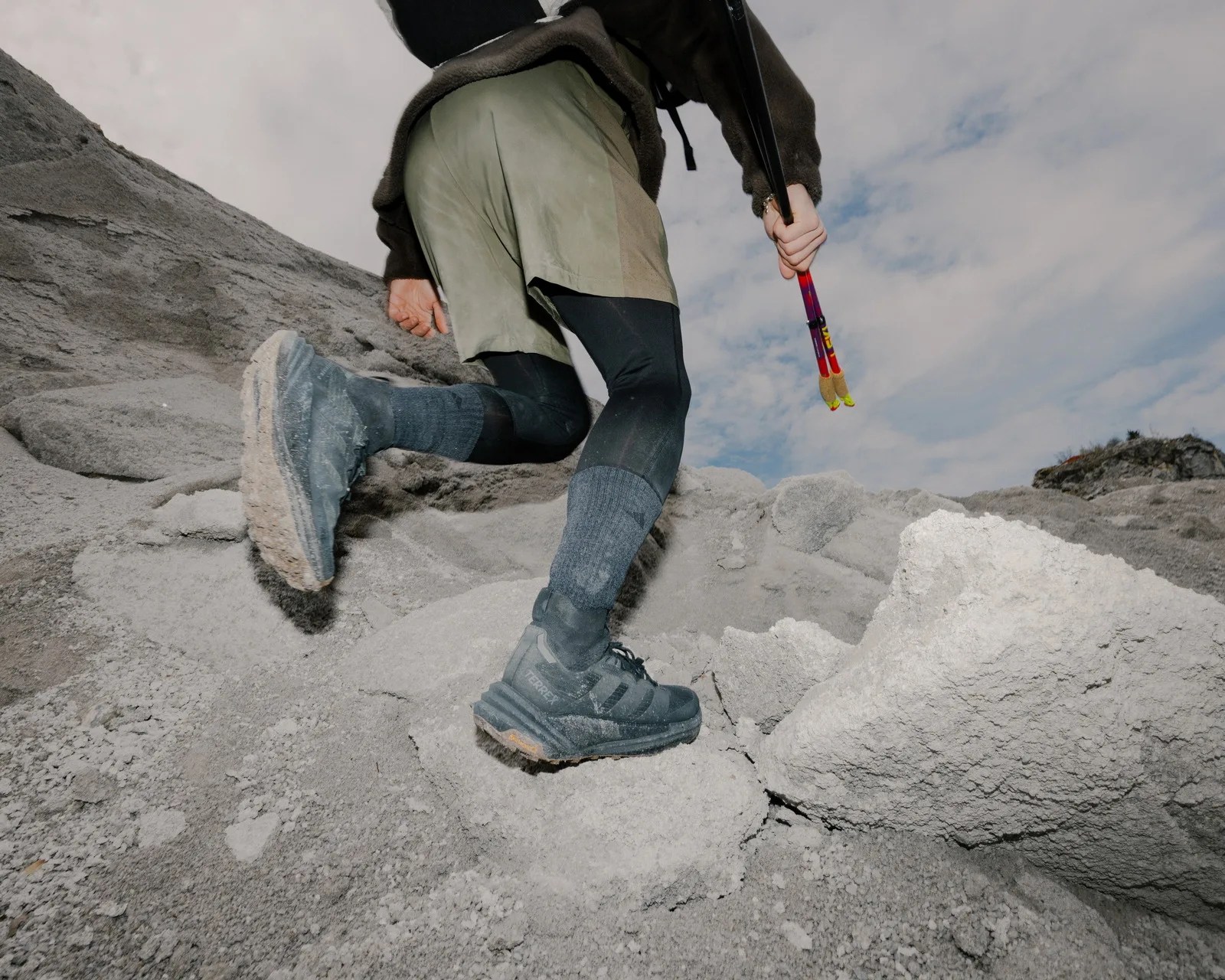 Person wearing black hiking shoes and black leggings climbing rocky terrain with trekking poles in hand.
