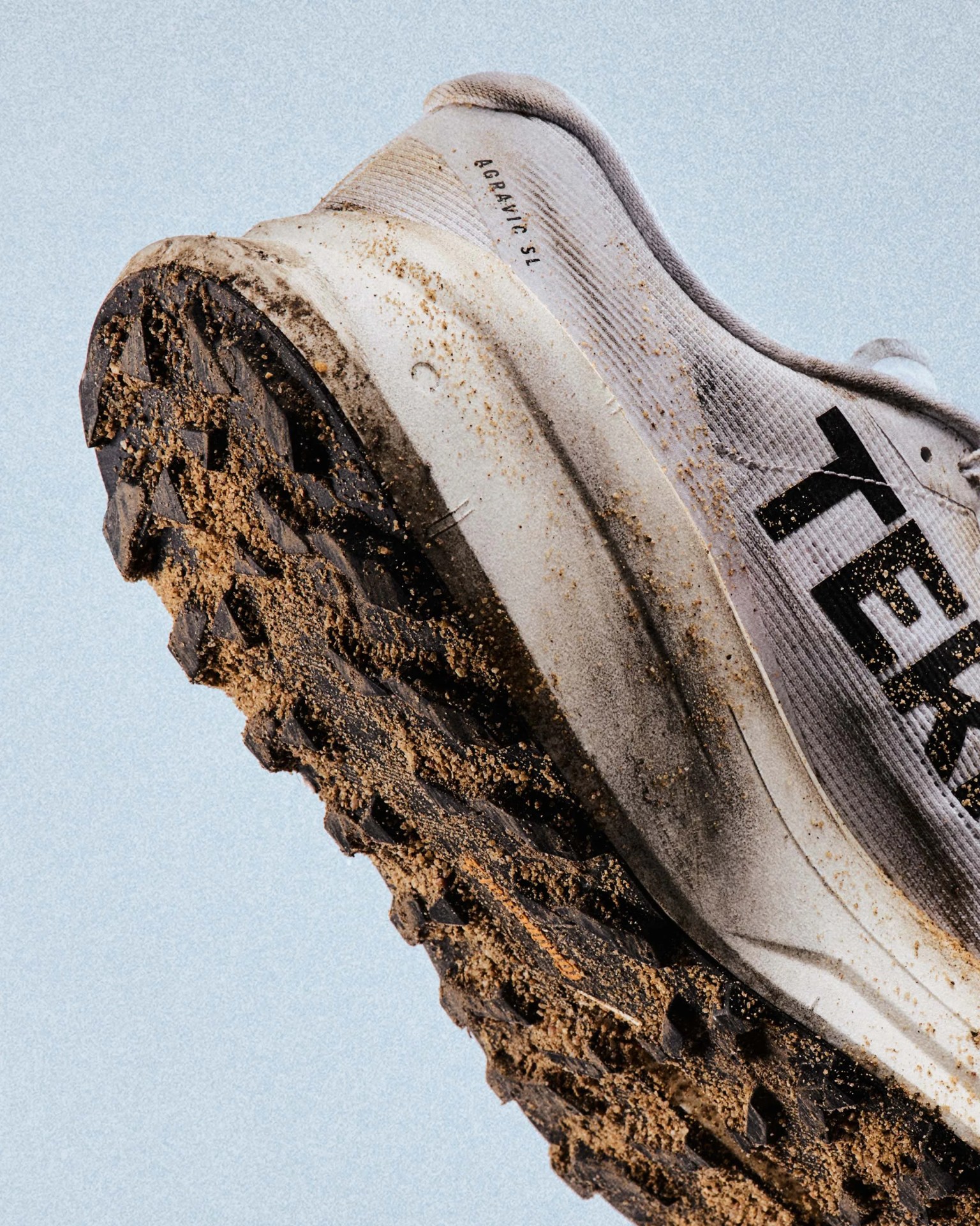White trail running shoe with black sole covered in dirt against a light blue background.