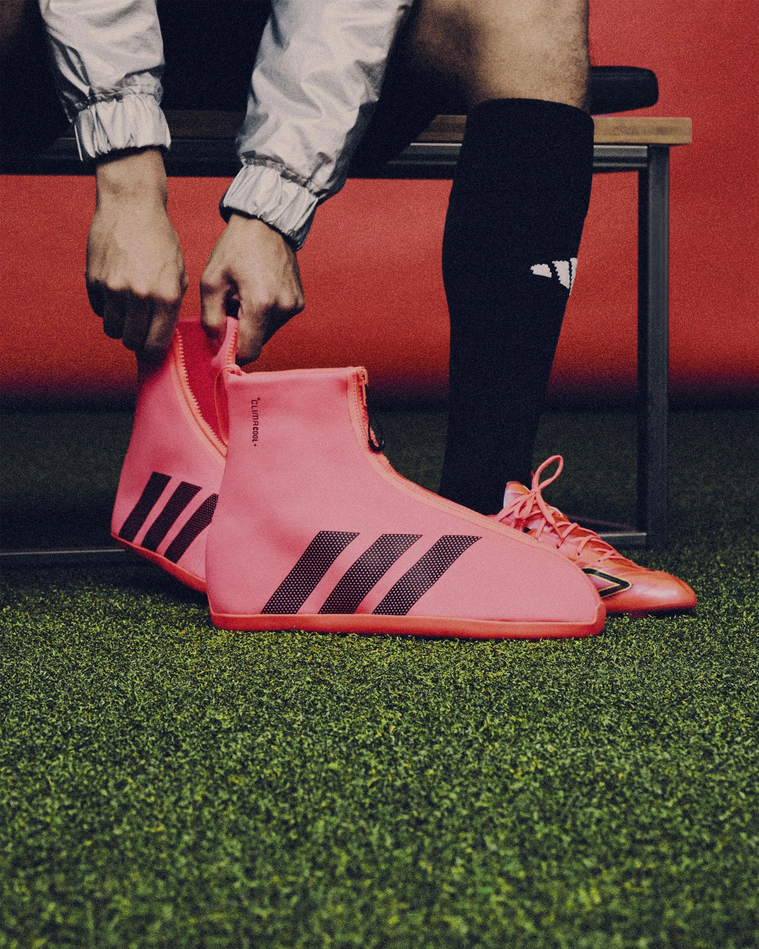 Person wearing black Adidas socks putting on bright pink Adidas soccer cleats with black stripes on artificial turf.