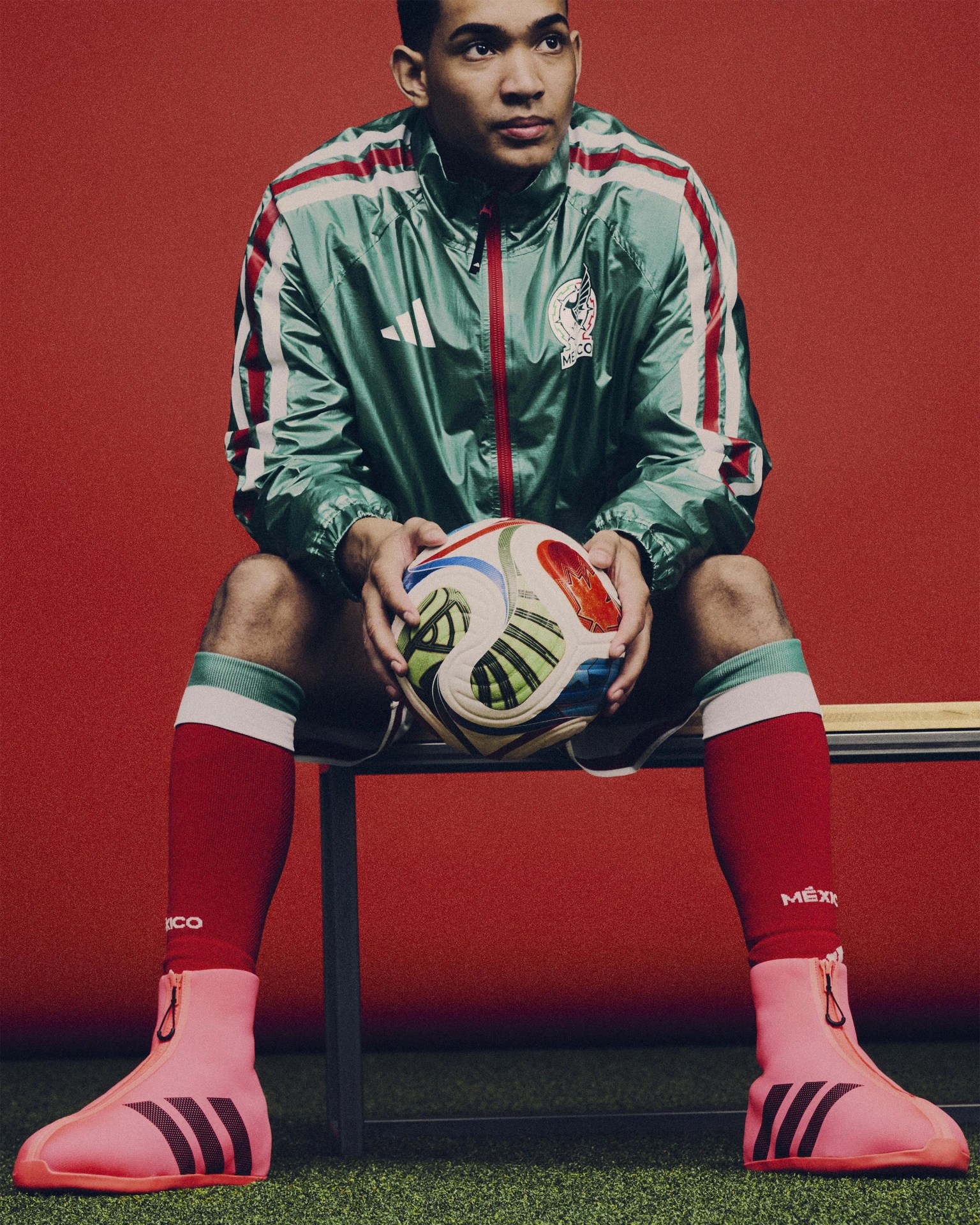 Person wearing a green Mexico soccer jacket, red socks, and pink Adidas soccer shoe covers holding a soccer ball.