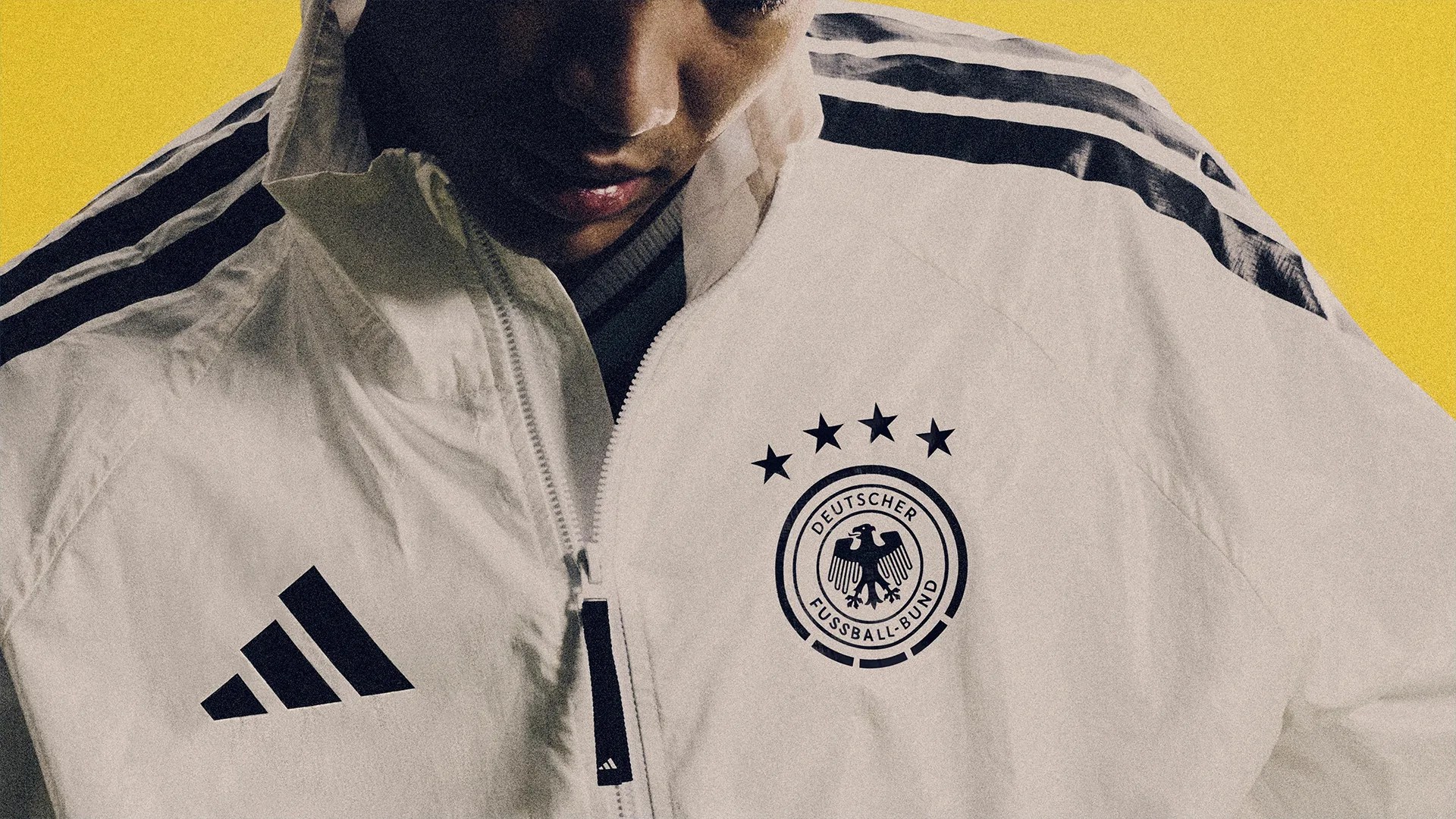 White Adidas jacket with black stripes and Deutscher Fussball-Bund logo worn by a person against a yellow background.