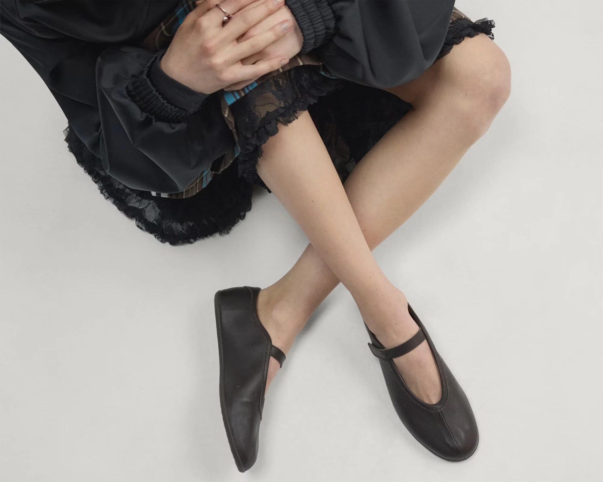 Person wearing black Mary Jane-style shoes with a strap, black lace-trimmed skirt, and black jacket, sitting cross-legged.