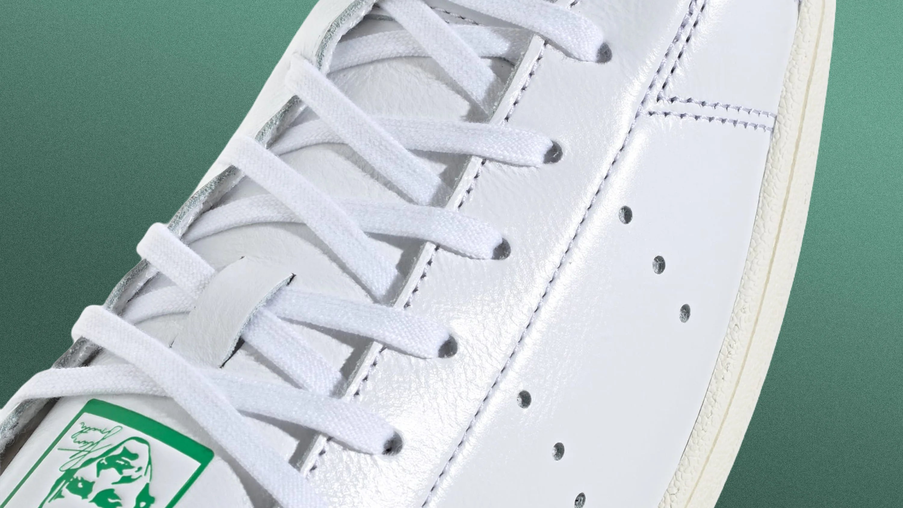 Close-up of white leather sneaker with white laces and green logo on tongue against green background.
