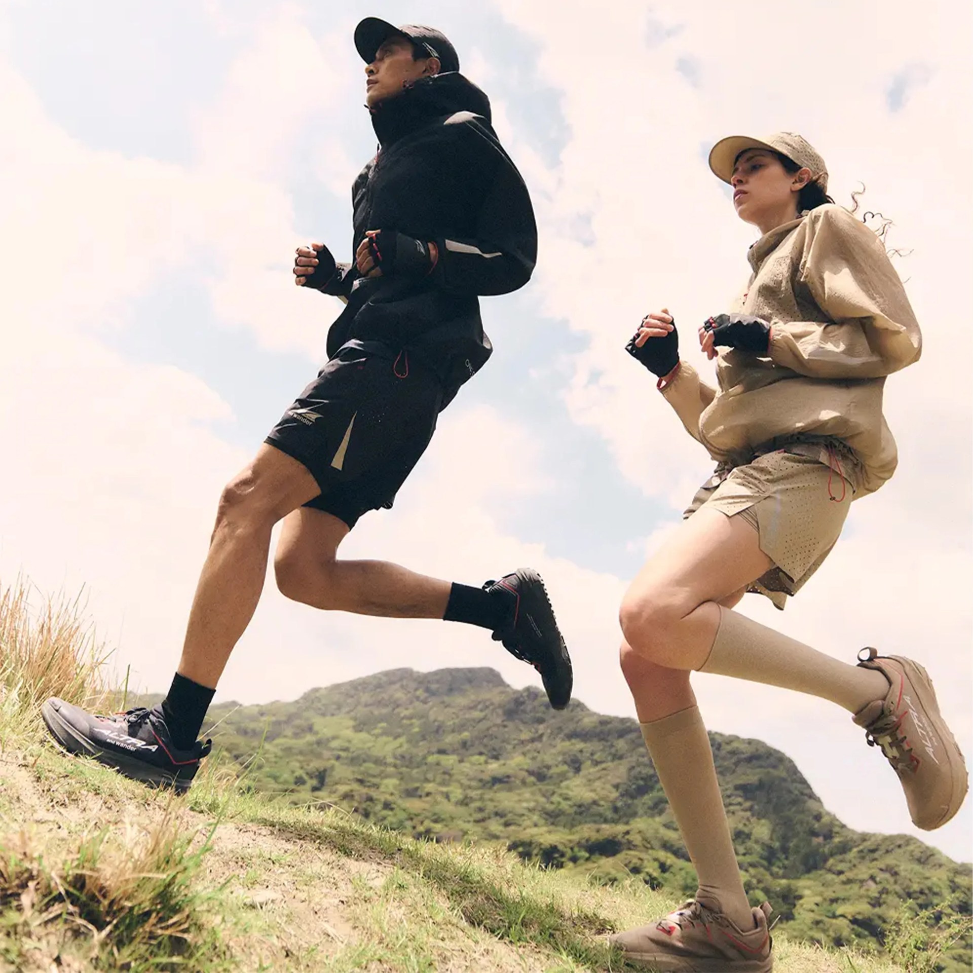 Two people running uphill wearing outdoor athletic clothing and trail running shoes against a cloudy sky.