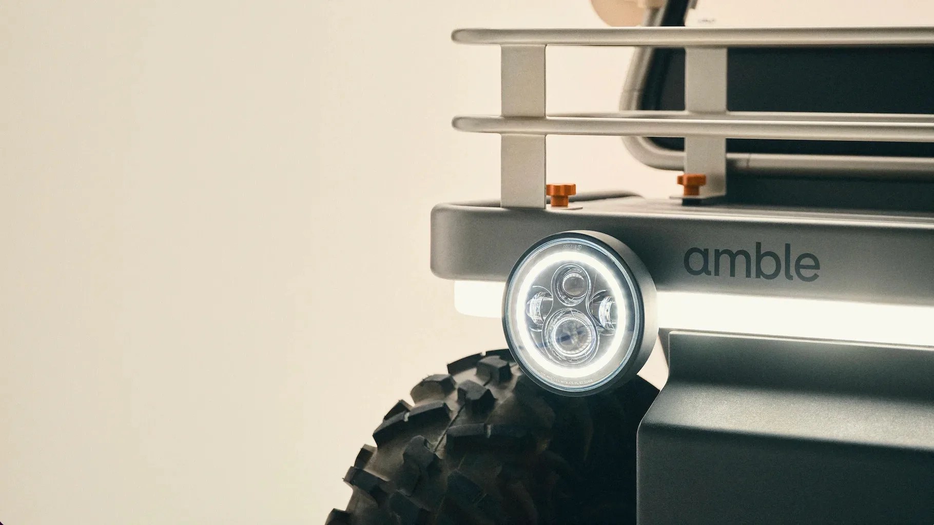 Close-up of a round LED headlight and rugged tire on a gray vehicle with "amble" branding.