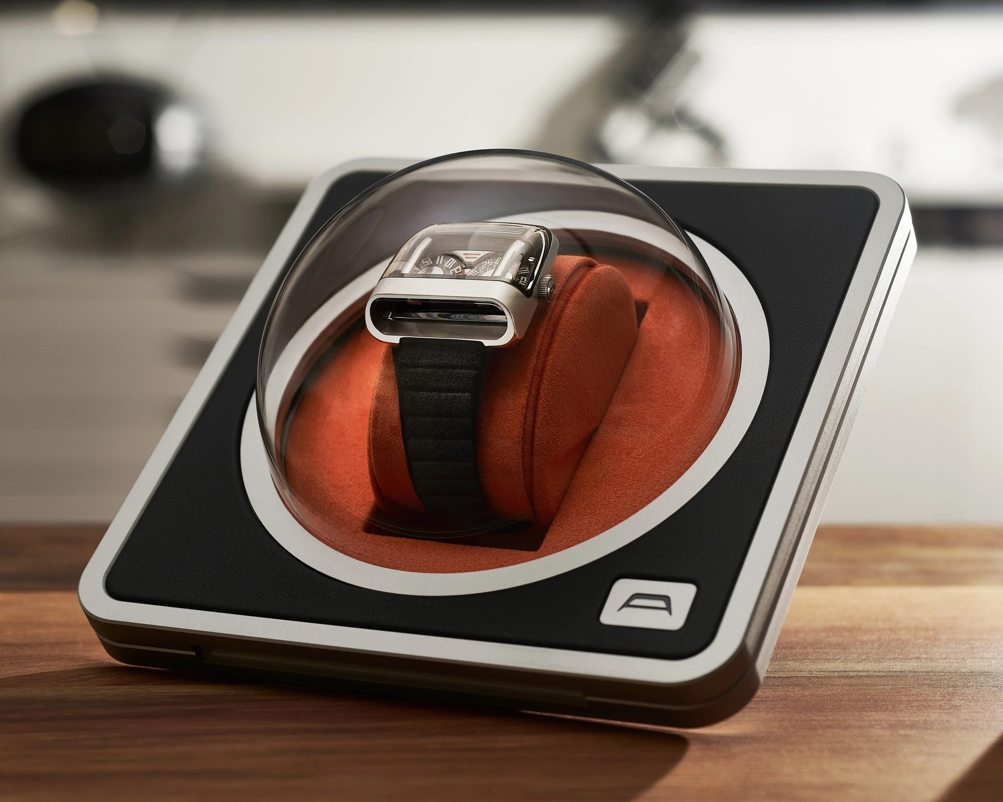 Silver futuristic wristwatch with black strap inside a clear dome on a black and silver display stand with orange cushion.