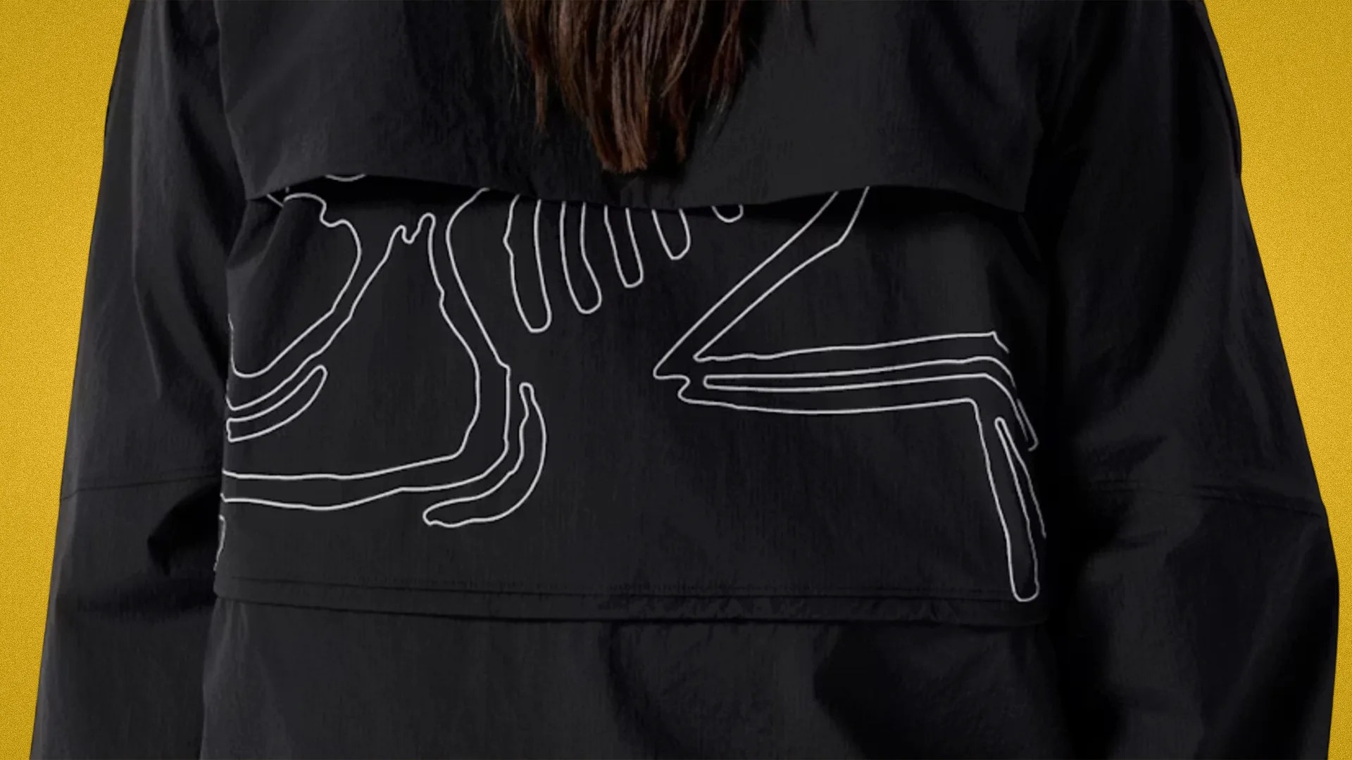 Black jacket with white abstract line design on the back, worn by a person against a yellow background.