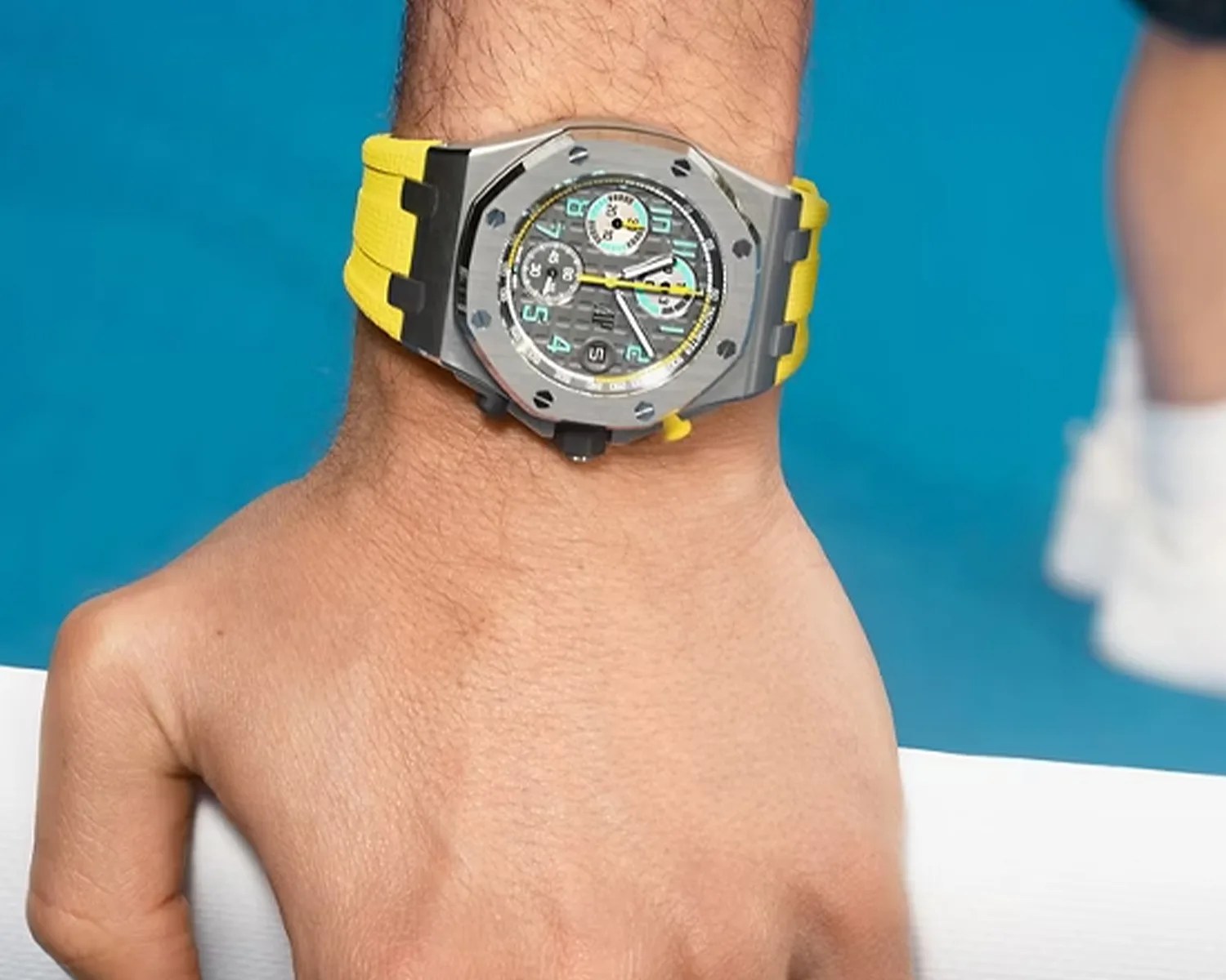 Silver octagonal wristwatch with black and yellow details and a yellow rubber strap worn on a wrist.
