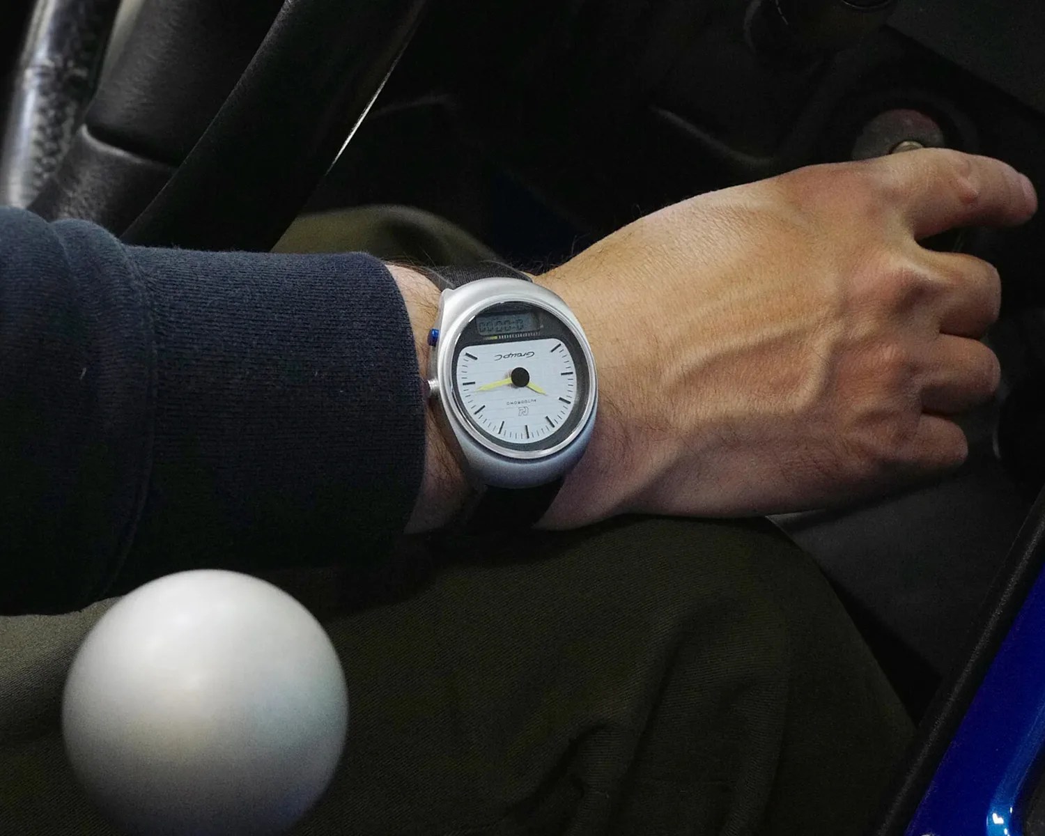 Silver analog-digital wristwatch with white face and black strap worn on a wrist near a car gear shift knob.
