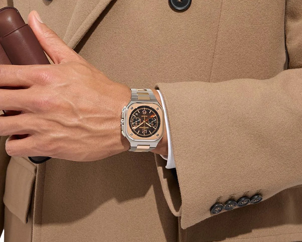 Silver and rose gold wristwatch with a black dial and chronograph on a wrist wearing a camel coat.
