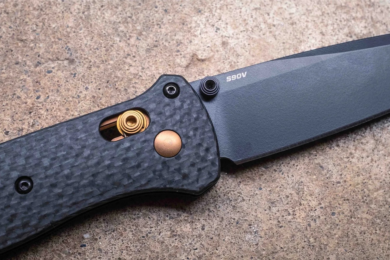 Black folding knife with textured carbon fiber handle and visible gold-colored spring mechanism on a stone surface.