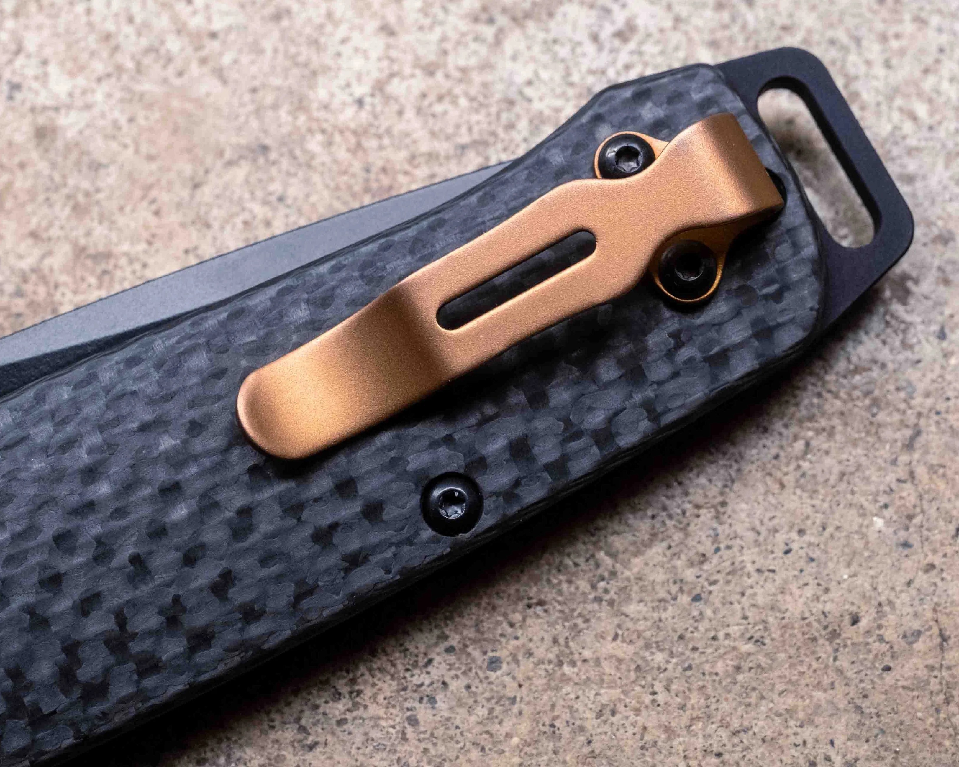 Close-up of a folding knife handle with a textured black carbon fiber grip, a bronze-colored metal pocket clip attached with two screws, and part of a black blade marked "S90V" visible. The background is a rough, light brown surface.