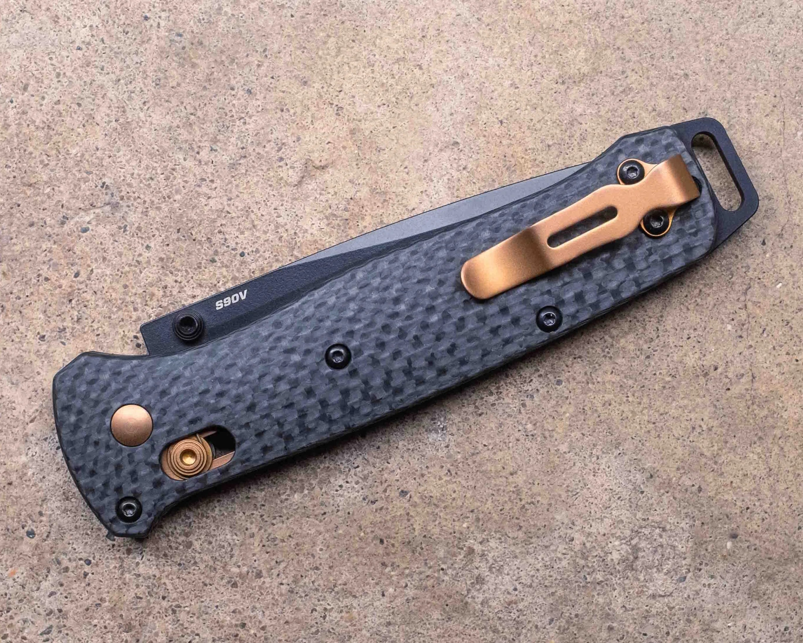 Folded pocket knife with a textured black handle, featuring a copper-colored clip and accents, resting on a rough beige surface. The blade is partially visible with the marking "A065.
