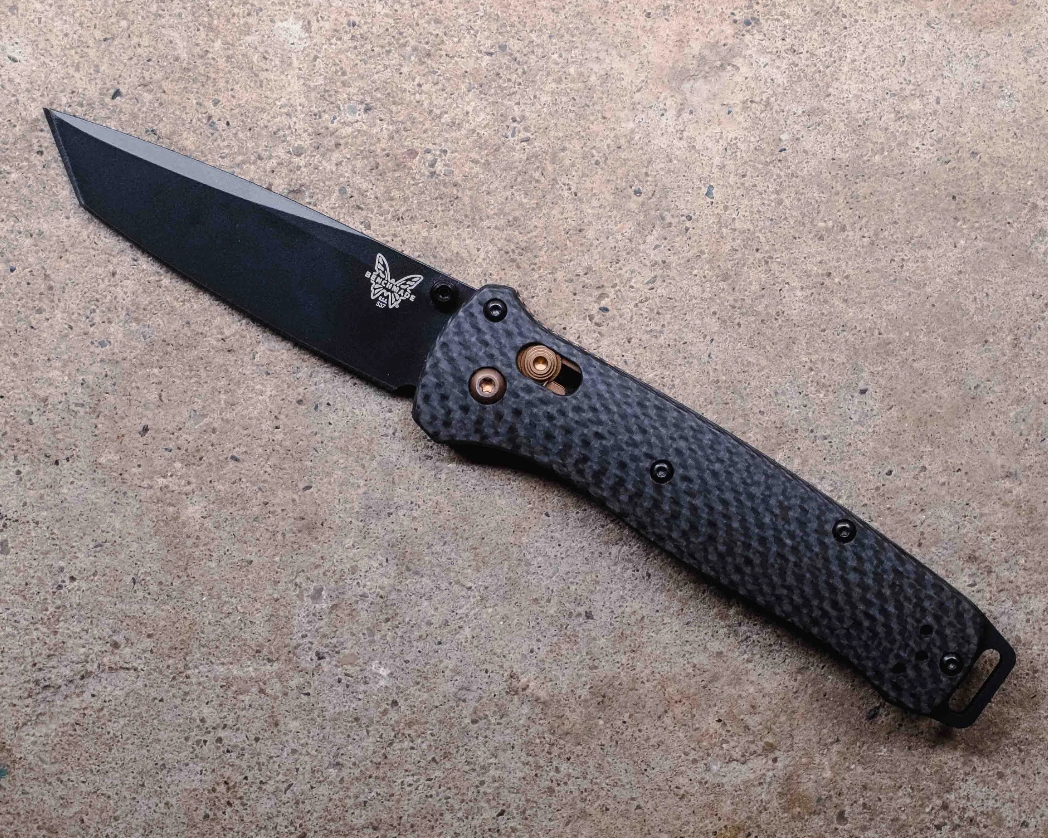Black folding knife with a textured handle and a tanto-style blade, featuring a butterfly logo near the base of the blade, placed on a rough, light brown concrete surface.