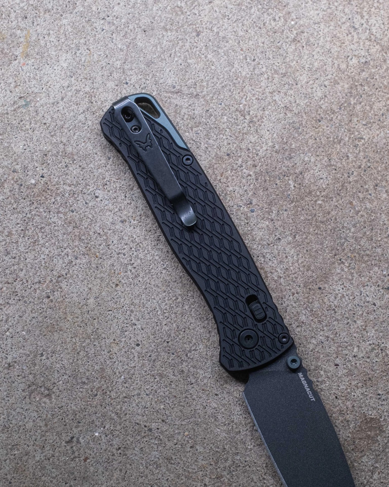 Black textured folding knife handle with metal clip and partially visible blade on concrete surface.