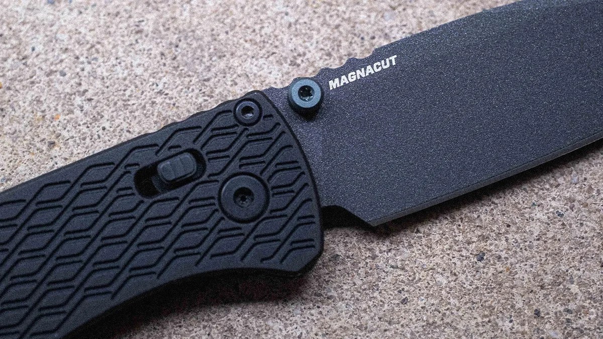 Black folding knife with textured handle and "MAGNACUT" printed on the blade, resting on a rough surface.