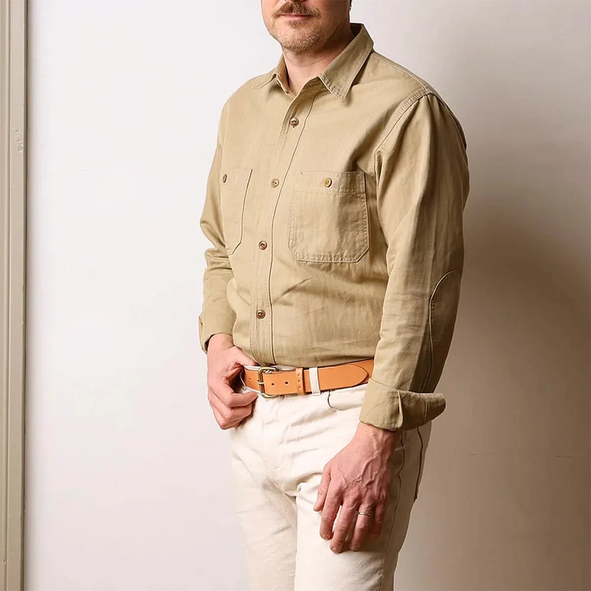 Man wearing a tan button-up shirt with two chest pockets and a tan leather belt with white pants.