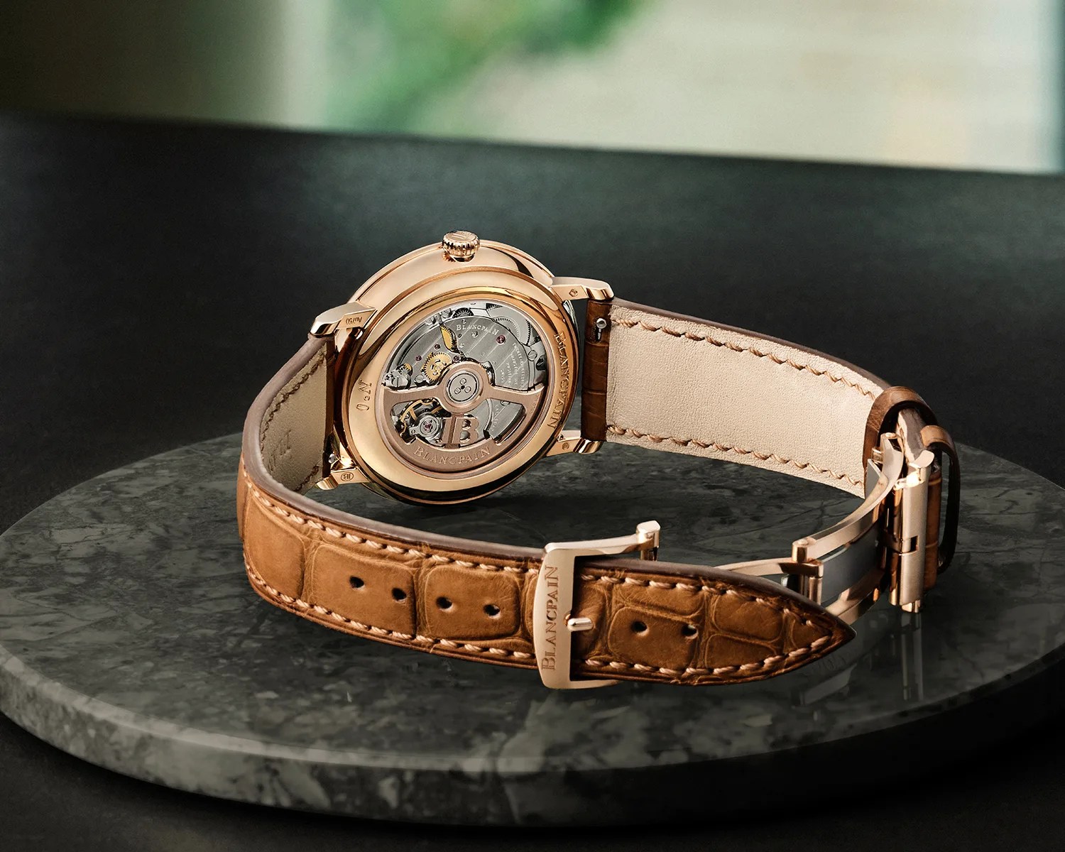 Rose gold Blancpain wristwatch with visible mechanical movement and brown leather strap on a marble surface.