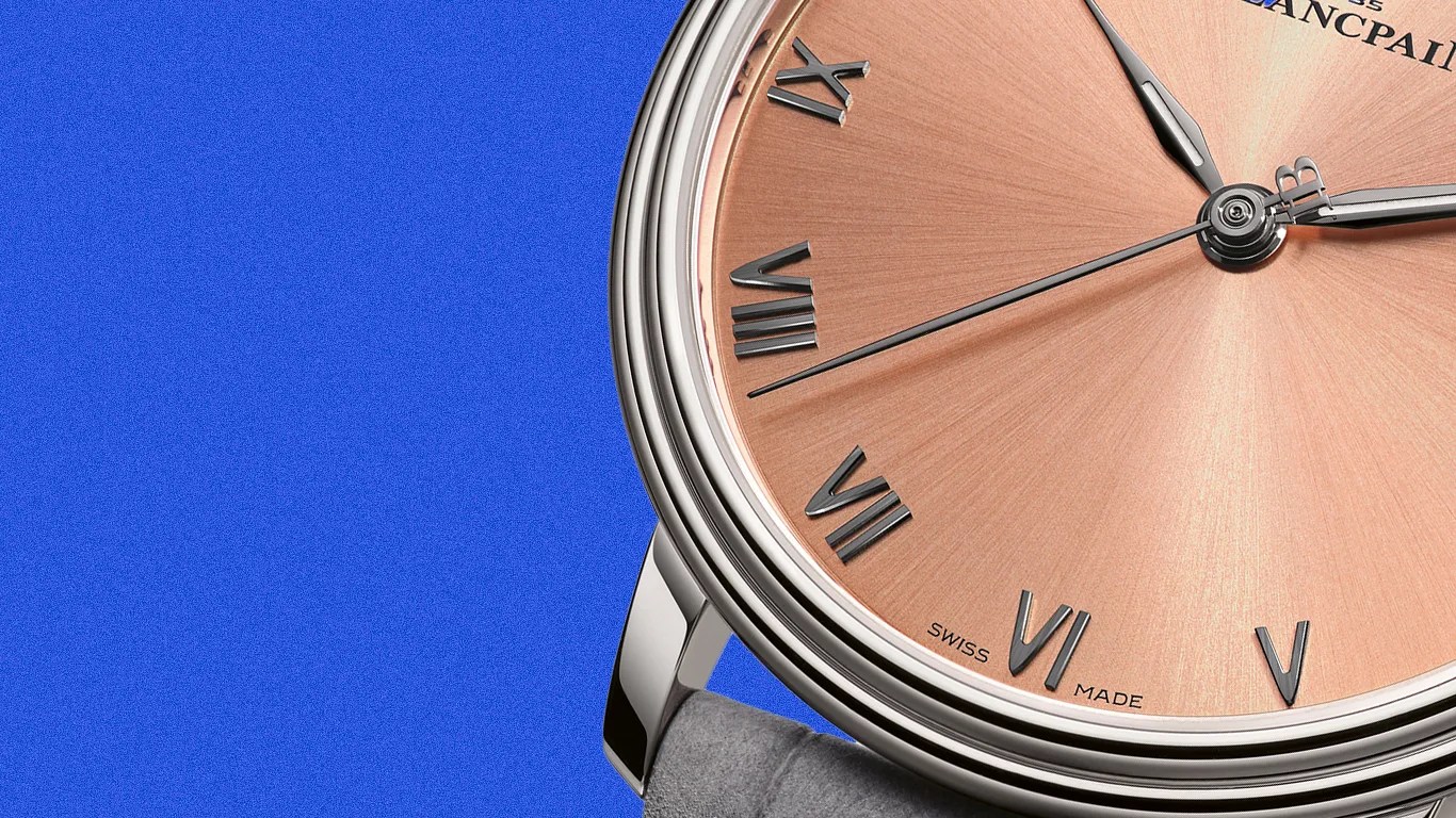 Close-up of a Blancpain watch with a rose gold dial, silver Roman numerals, and a gray leather strap against a blue background.