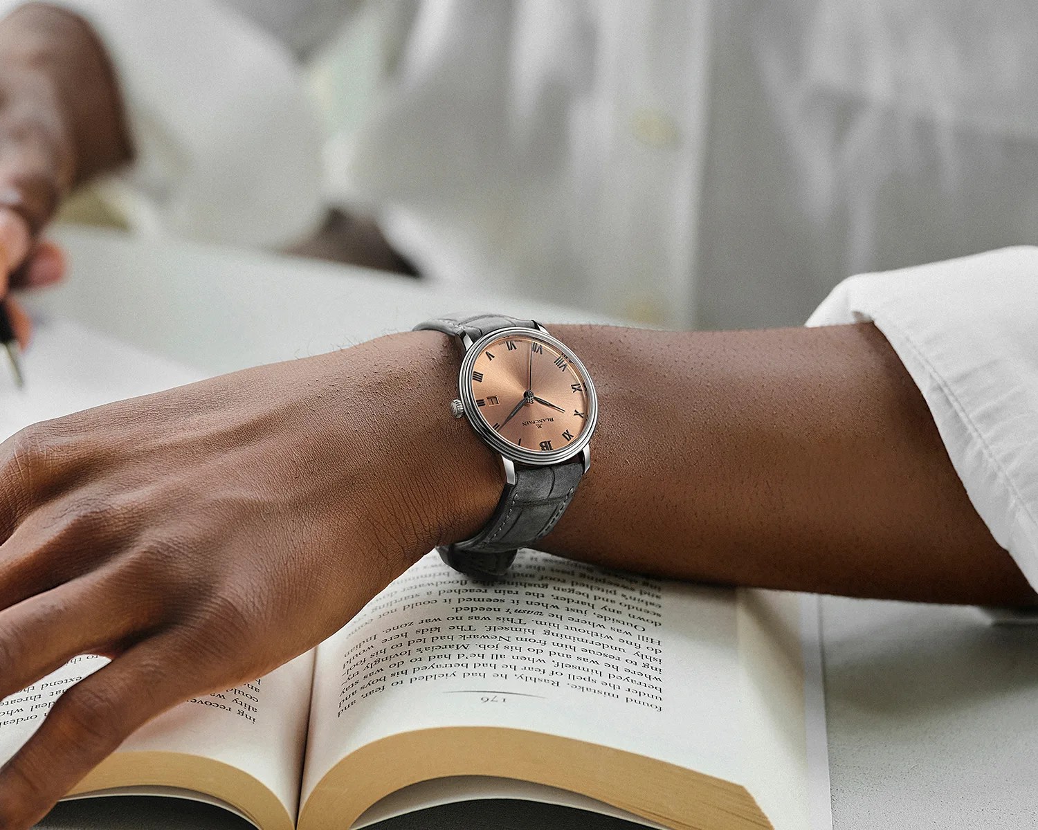 Silver wristwatch with a rose gold dial and black Roman numerals on a gray leather strap worn on a wrist over an open book.