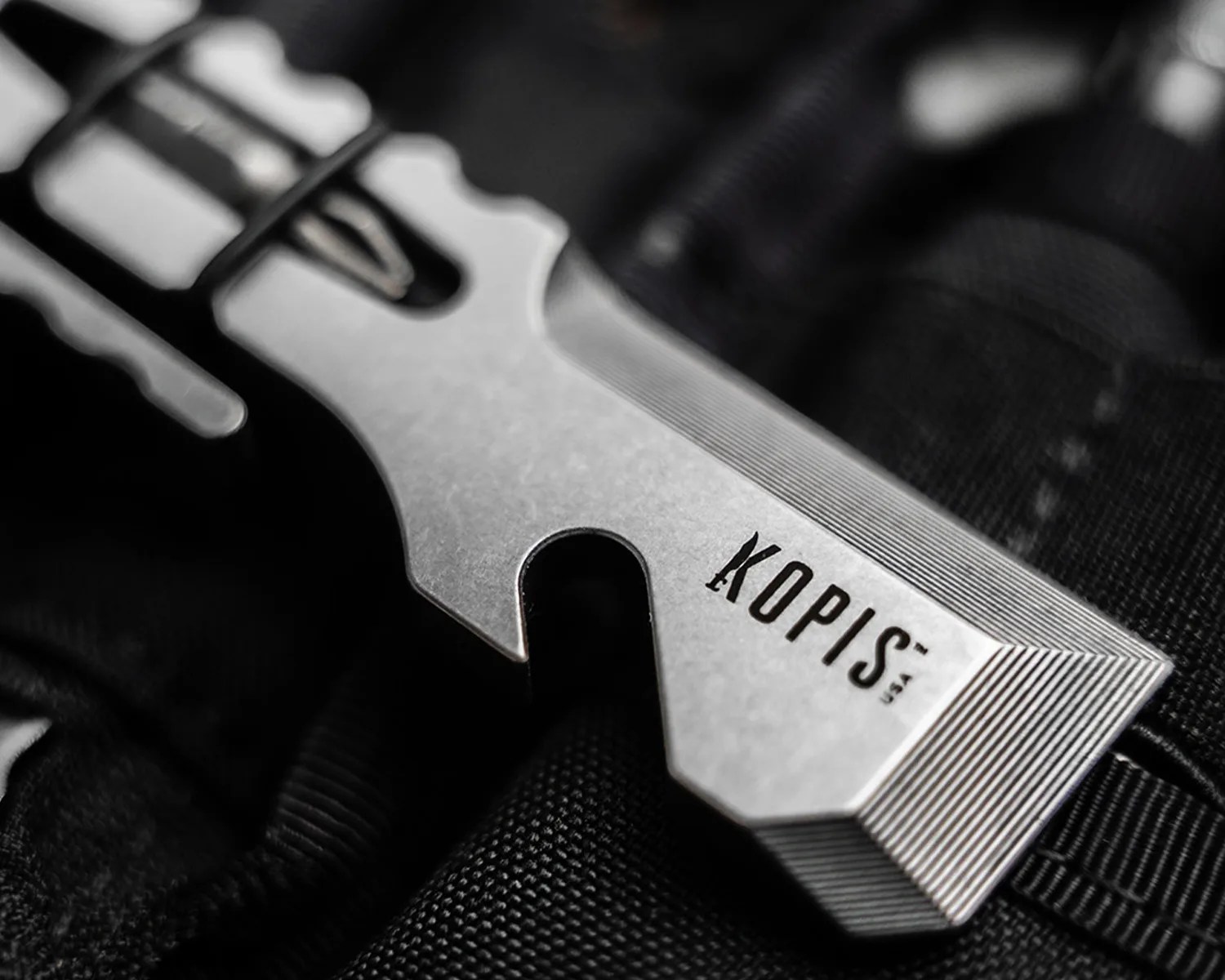 Close-up of a silver metal multitool with the word "KOPIS" engraved on it, resting on black fabric.
