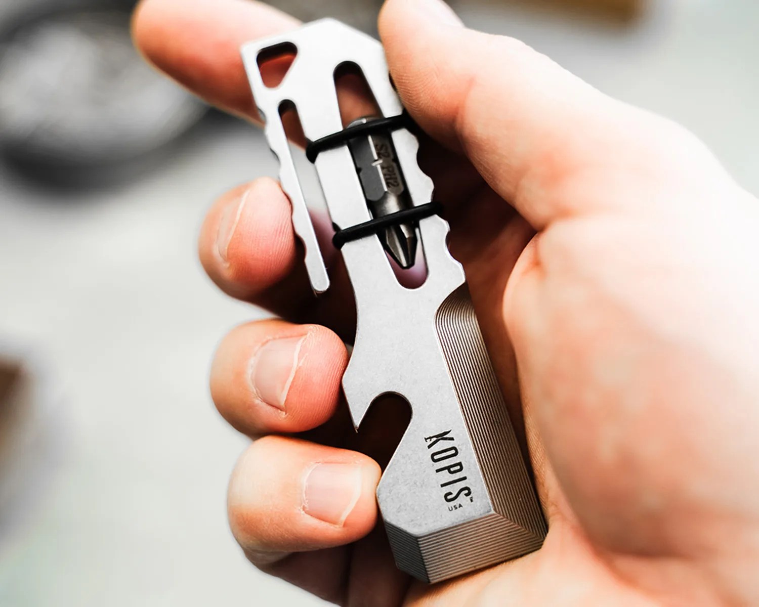 Hand holding a compact silver multi-tool with a black bit secured by rubber bands and "KOPIS USA" engraved.