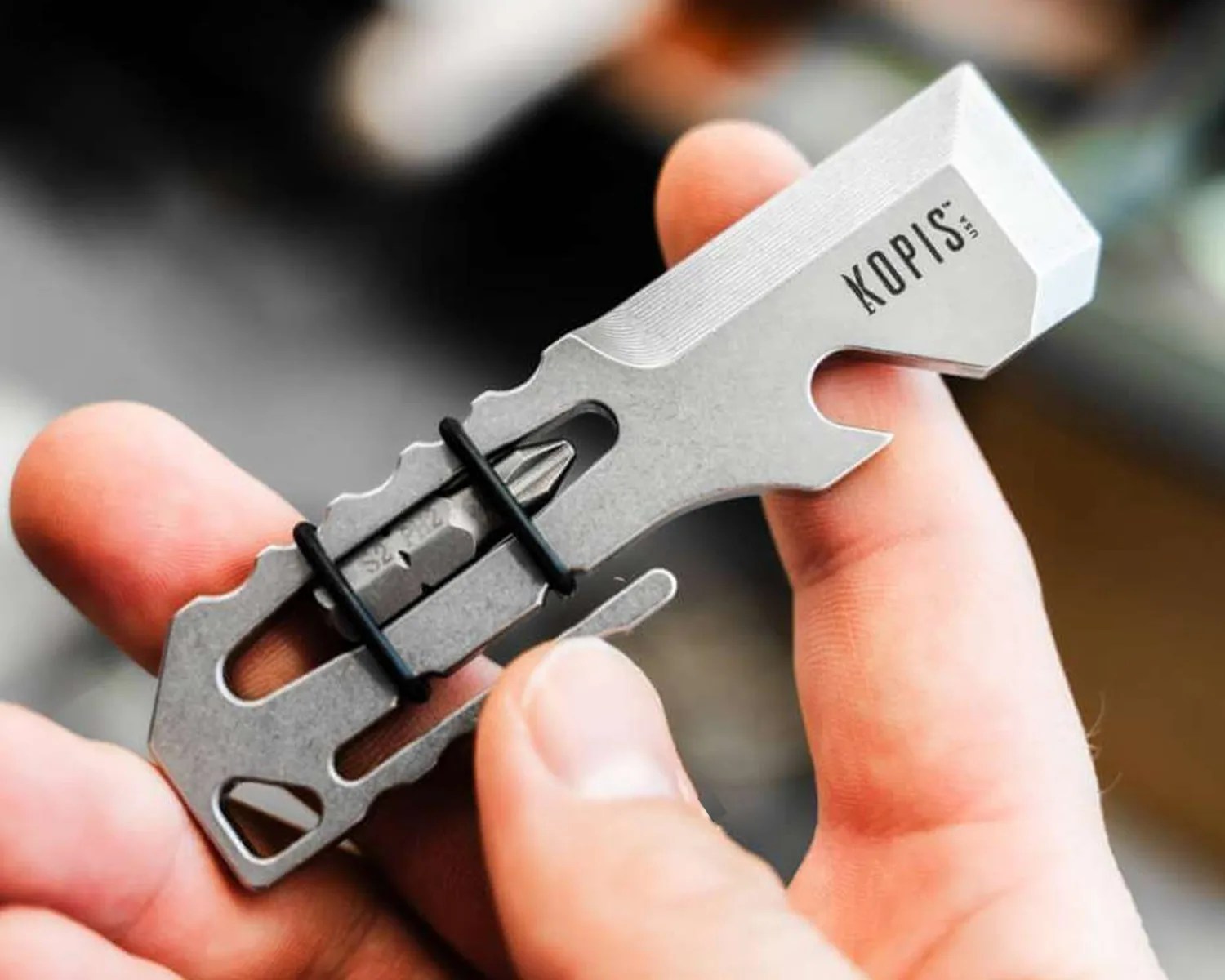 Compact silver multi-tool with screwdriver bit held between fingers, branded "KOPIS" on one end.