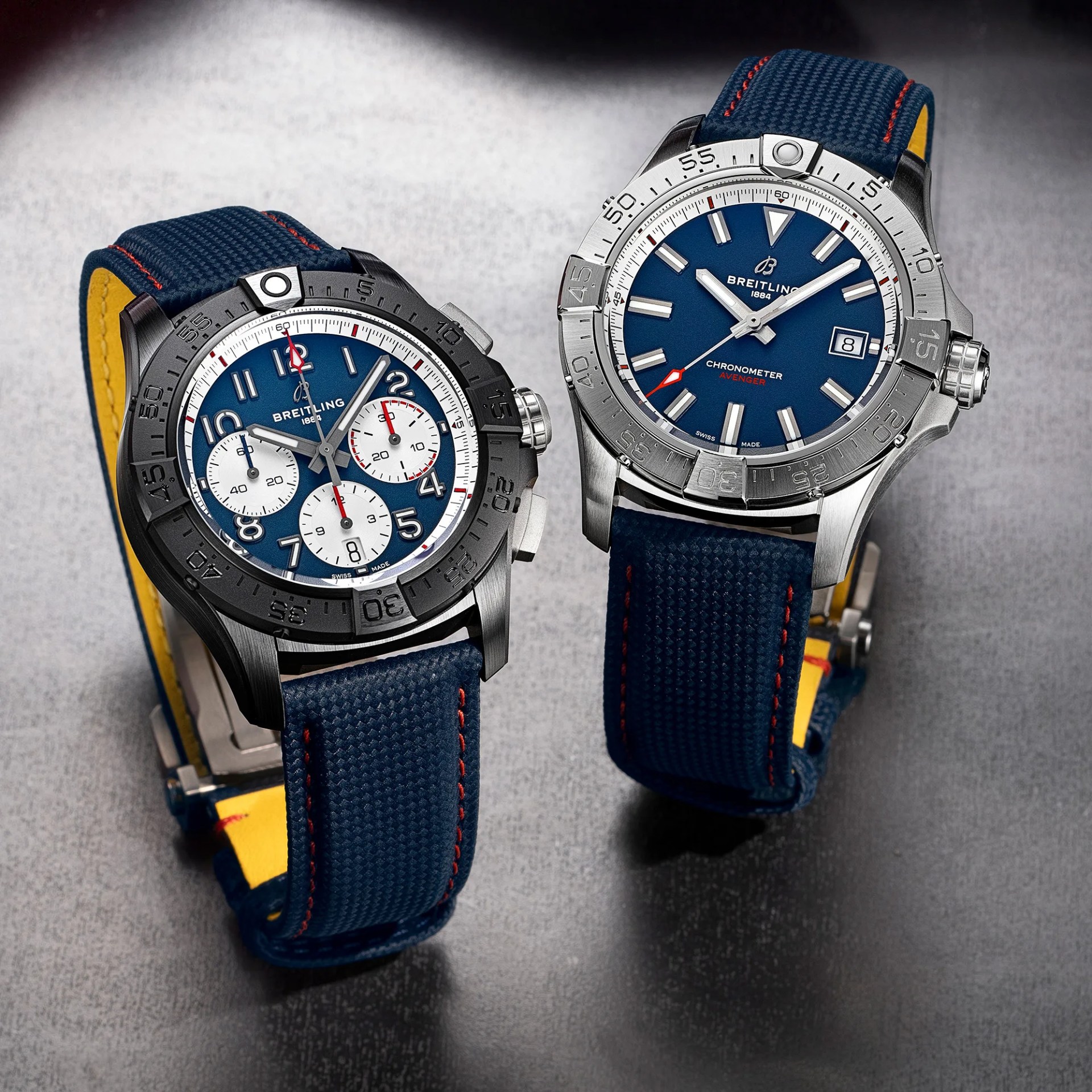 Two Breitling wristwatches with blue dials, silver bezels, and blue fabric straps with yellow undersides on a gray surface.