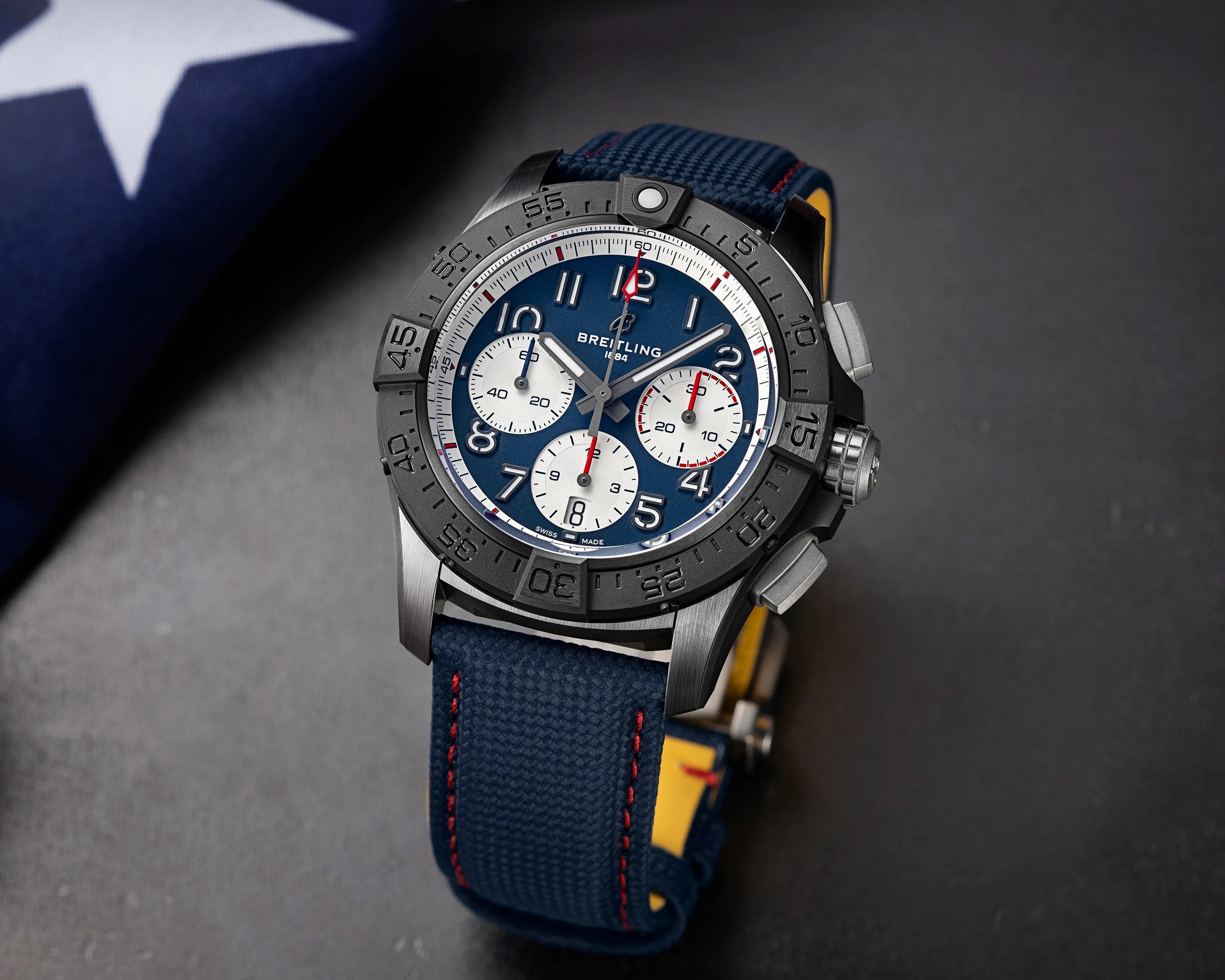 Breitling chronograph watch with blue dial, black bezel, and blue fabric strap with red stitching.