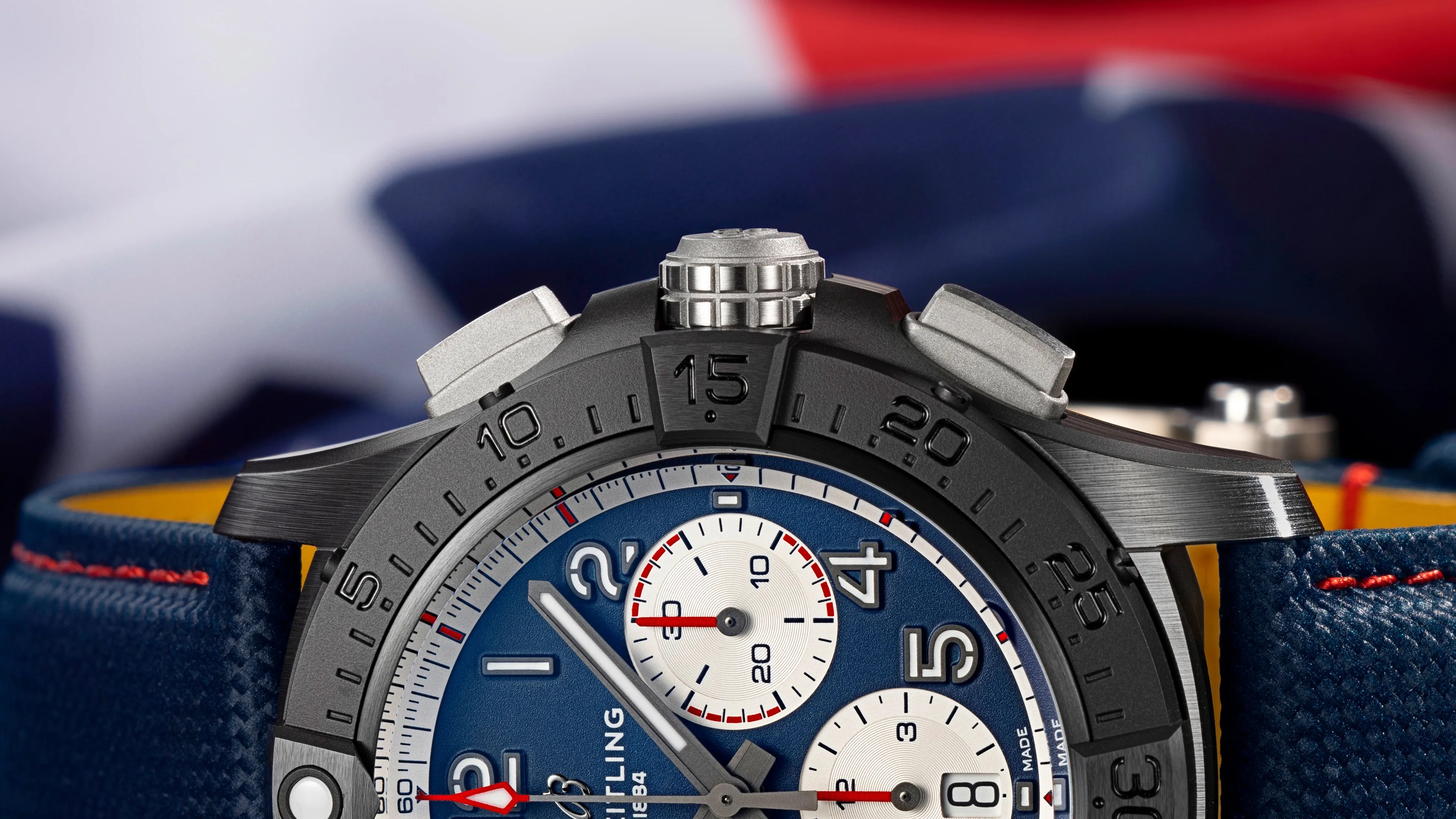 Close-up of a black bezel chronograph watch with blue dial, white subdials, and blue textured strap with red stitching.