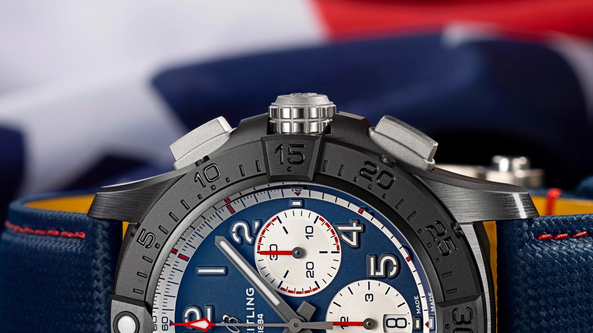 Close-up of a black bezel chronograph watch with blue dial, white subdials, and blue textured strap with red stitching.